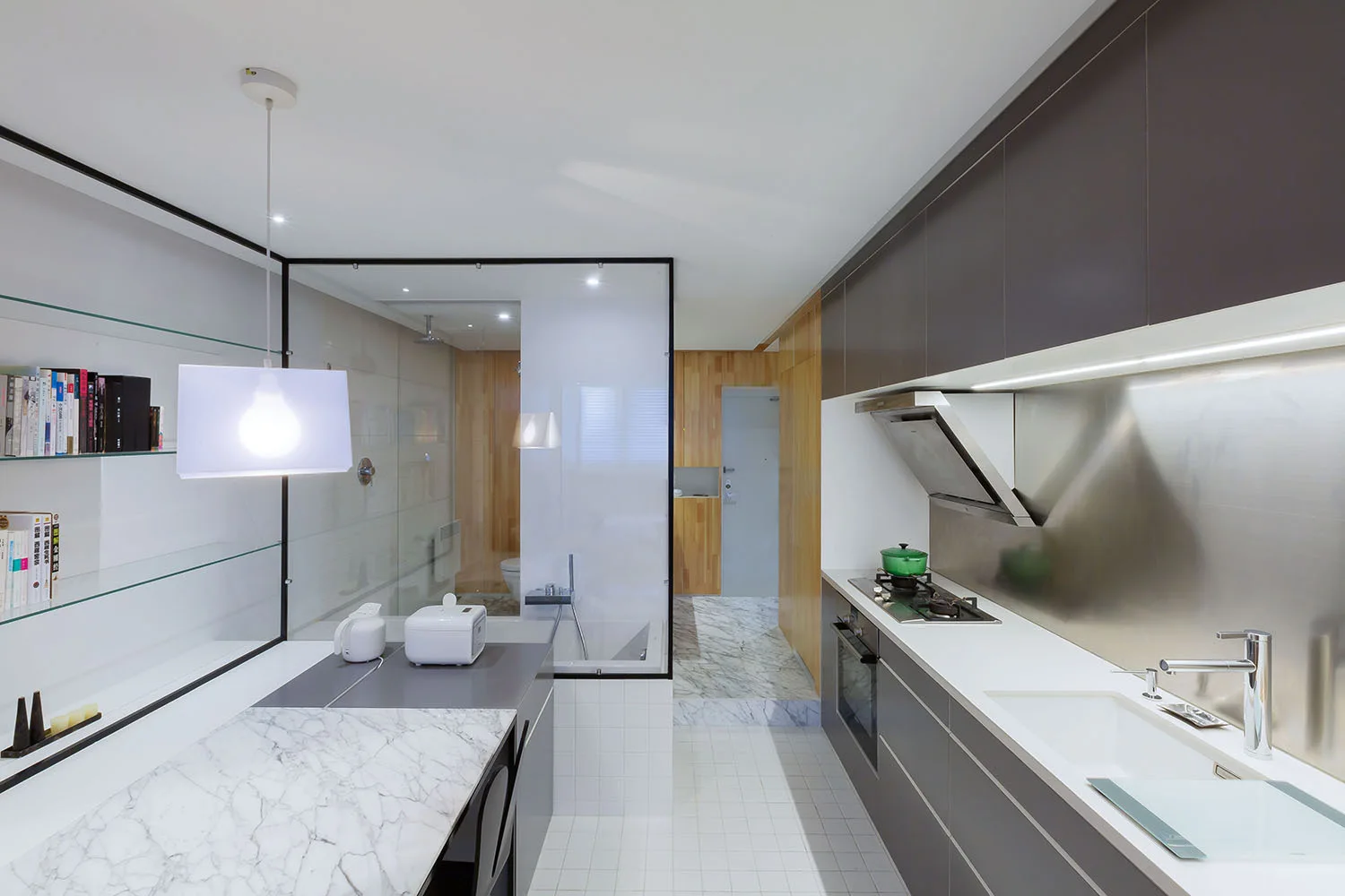 A modern kitchen with grey cabinetry, a marble-topped island, a stainless steel backsplash, and a black-framed glass partition wall.