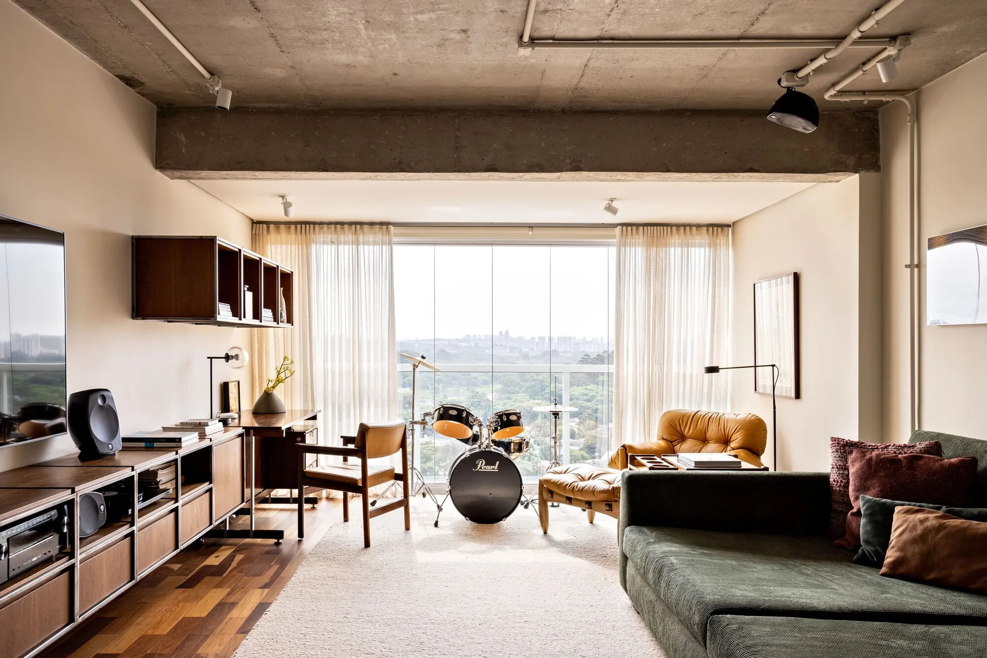 An industrial living room featuring an exposed concrete ceiling, a green sofa, custom wood cabinetry, and a drum kit in front of a window.