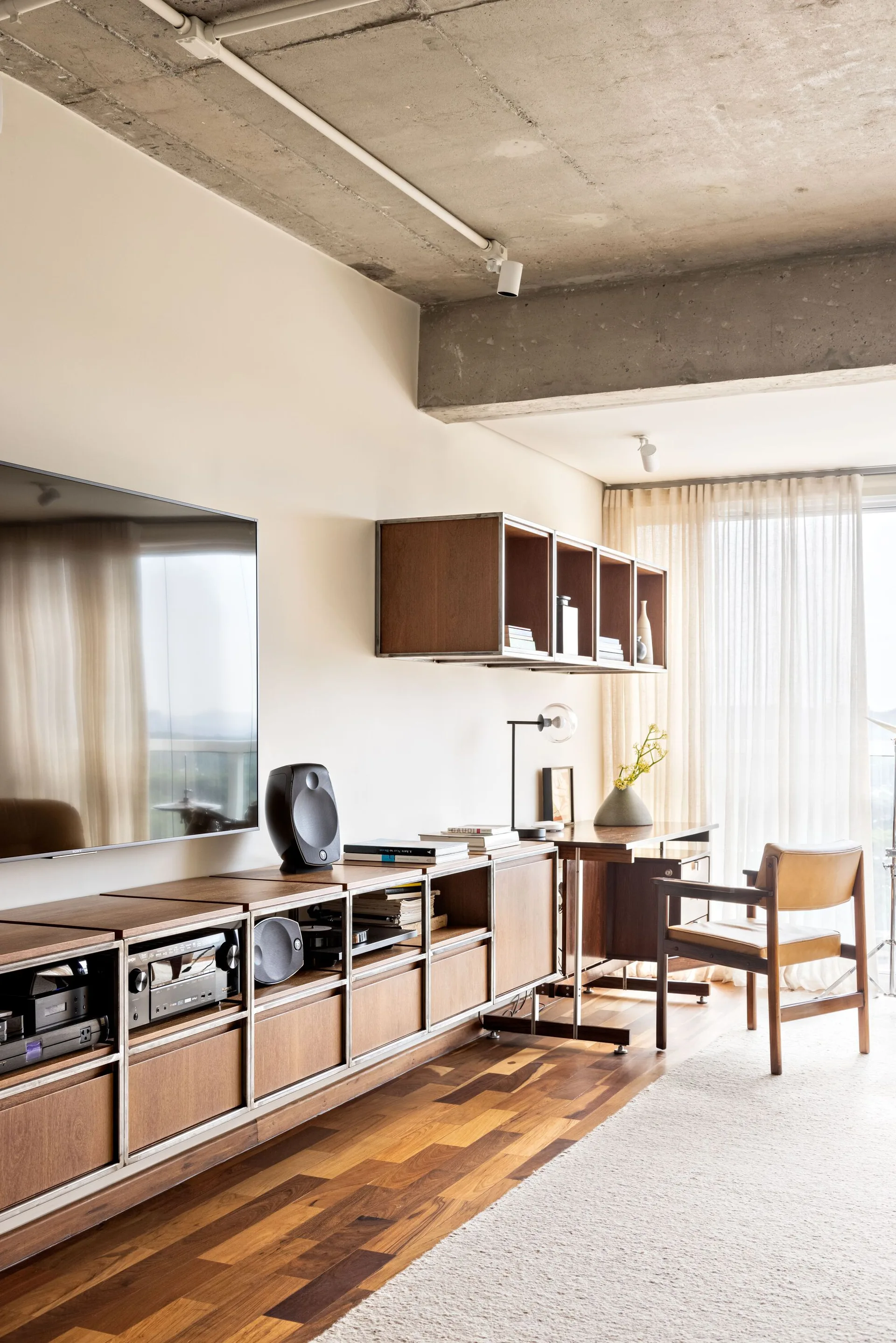 A long wooden media console with metal framing extends into a desk against an off-white wall under an exposed concrete ceiling.