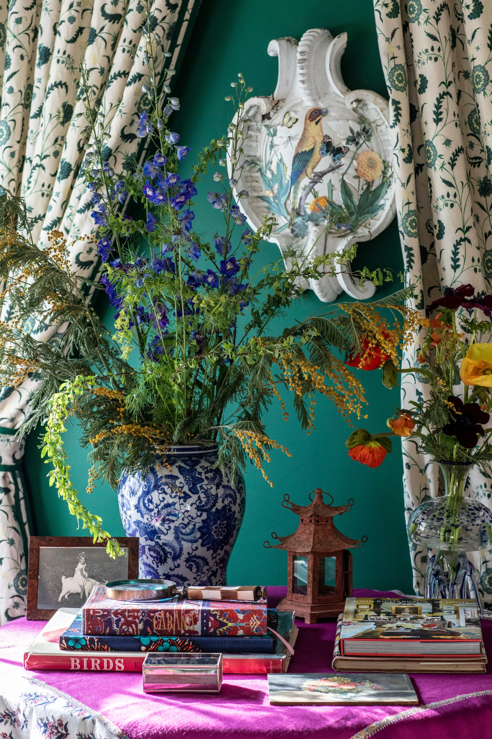 A tabletop vignette features a blue and white ceramic vase with tall blue flowers against a green wall flanked by botanical curtains.
