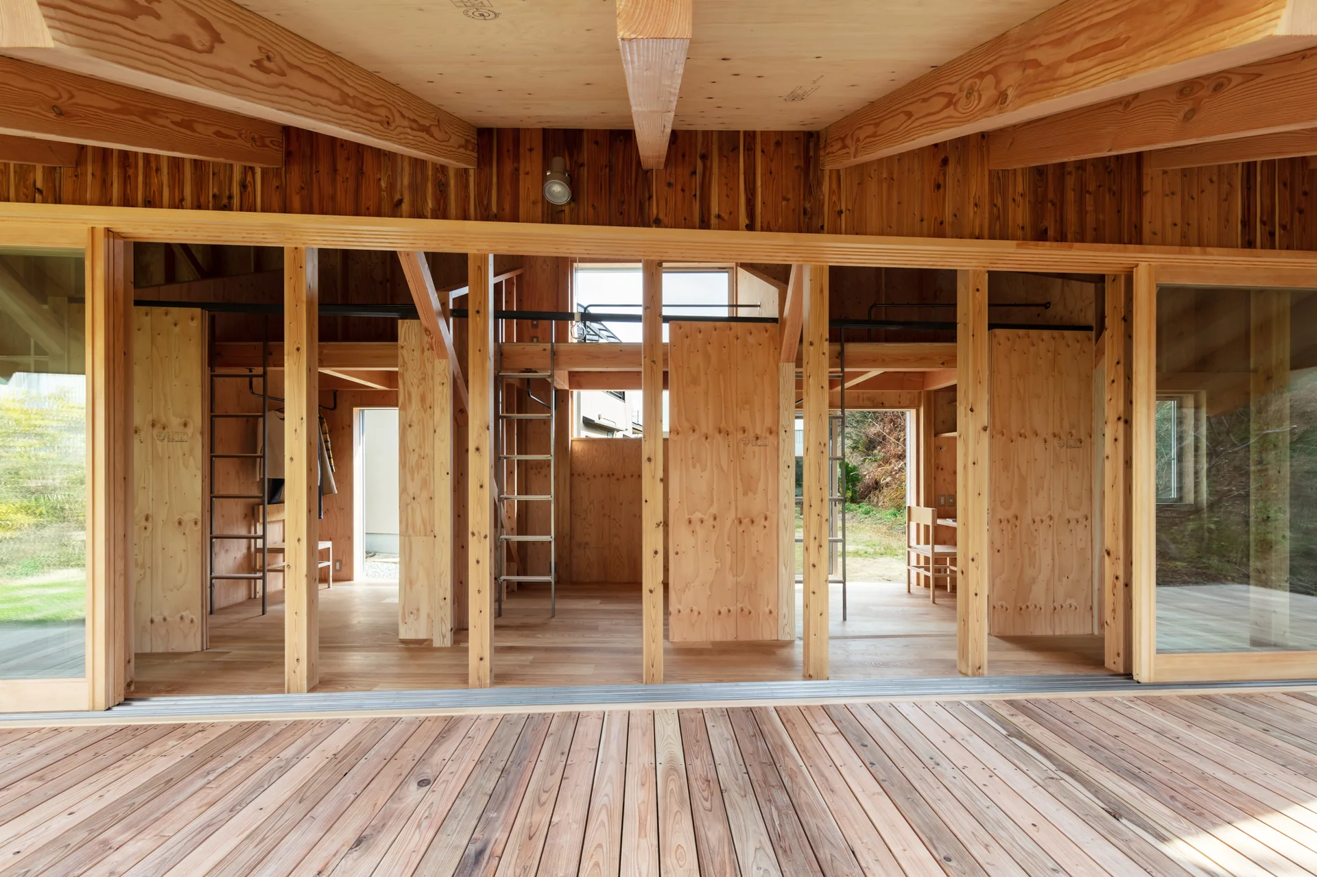 A minimalist wooden cabin interior with raw plywood walls, exposed ceiling beams, and sliding glass doors opening onto an outdoor deck.