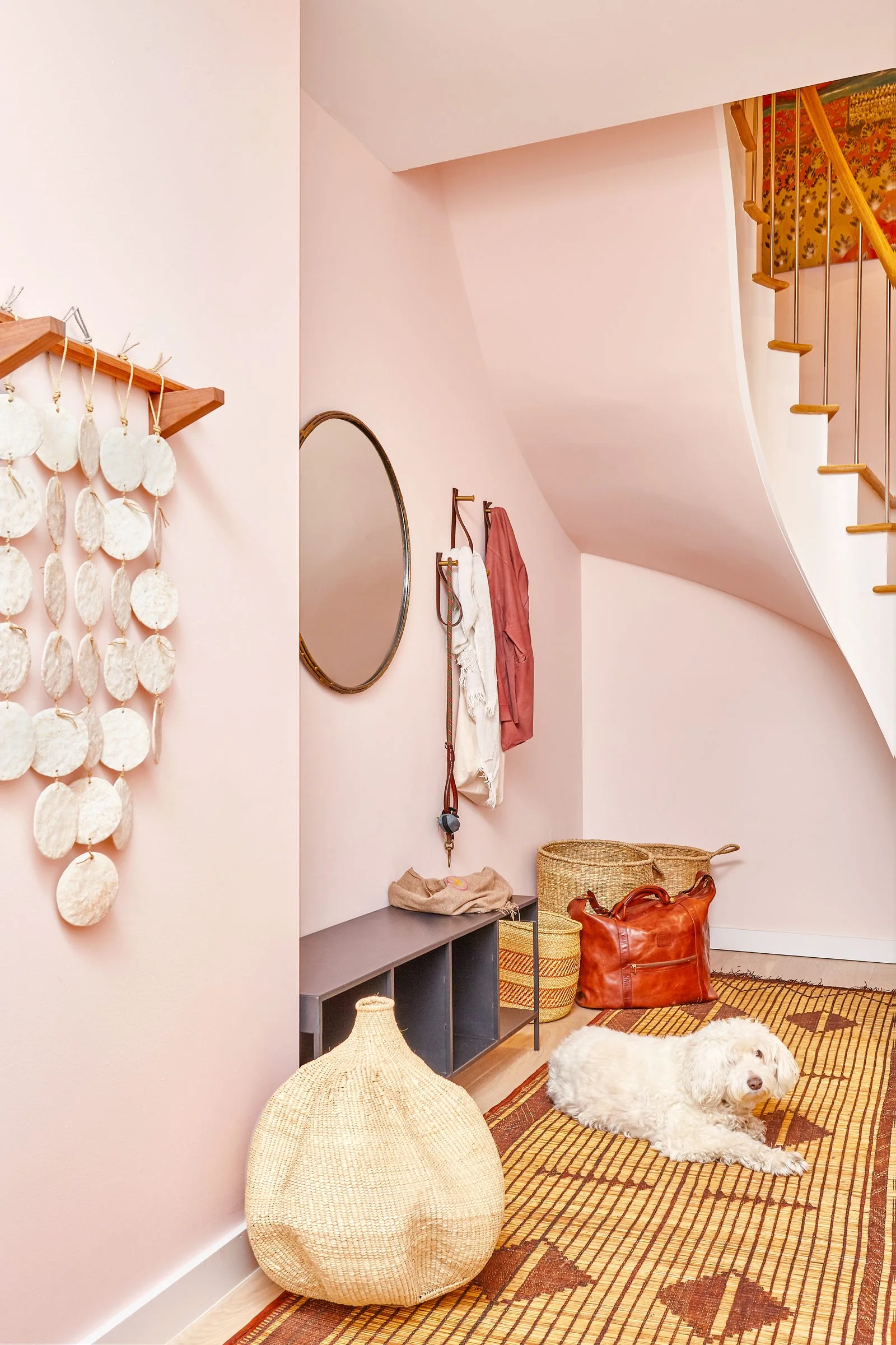 A pale pink entryway under a curved staircase features a grey storage bench, woven baskets, a round mirror, and a patterned fiber rug.