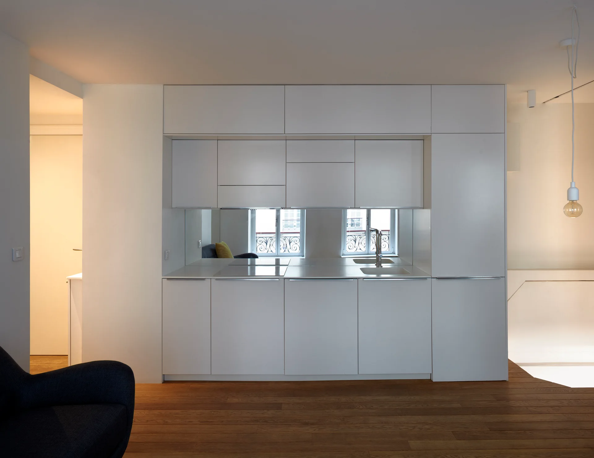 A minimalist white kitchen cabinet unit built into a wall featuring a mirrored backsplash, integrated sink, and dark wood flooring.
