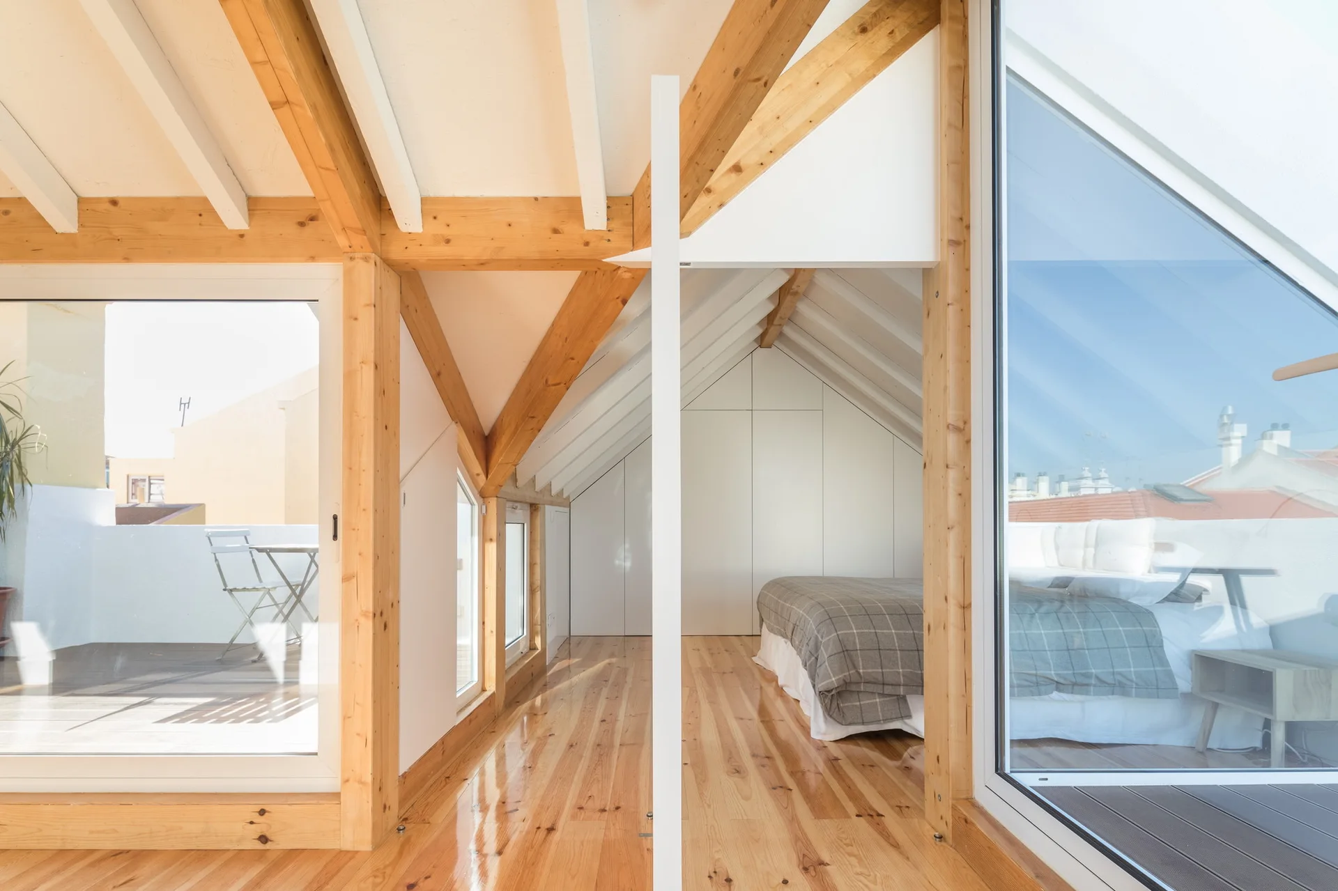 An attic bedroom with exposed wood ceiling beams, polished hardwood floors, and a glass partition looking into the bed area.