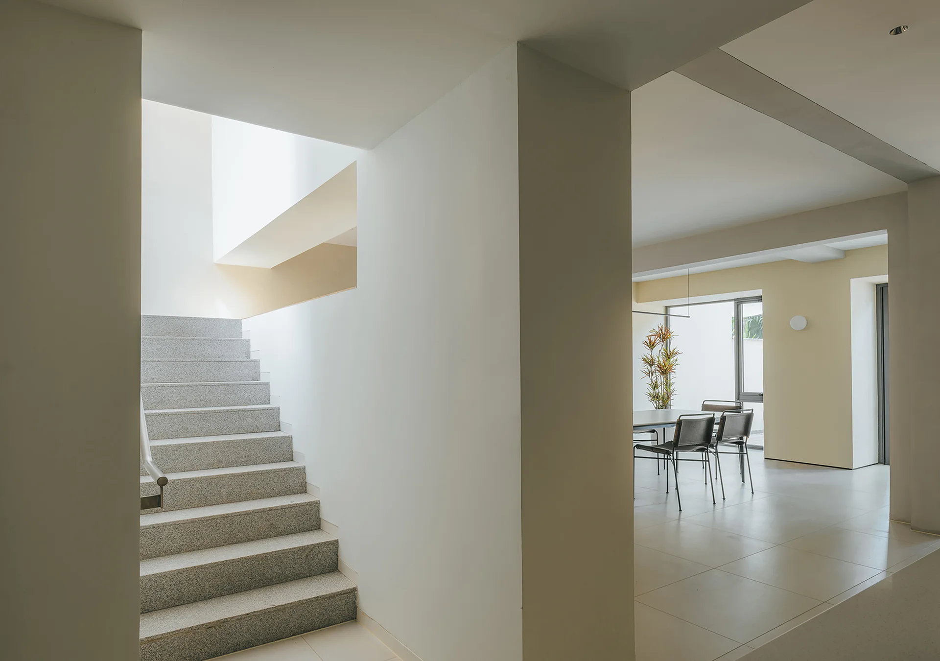 A minimalist interior features a speckled stone staircase next to an open-plan dining area with a modern table, black chairs, and beige tiled floors.