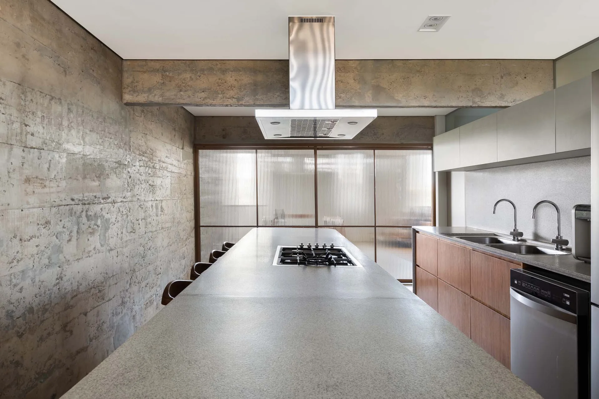 A modern kitchen features a long concrete island with a cooktop, raw concrete walls, warm wood lower cabinets, and ribbed glass sliding doors in the back.