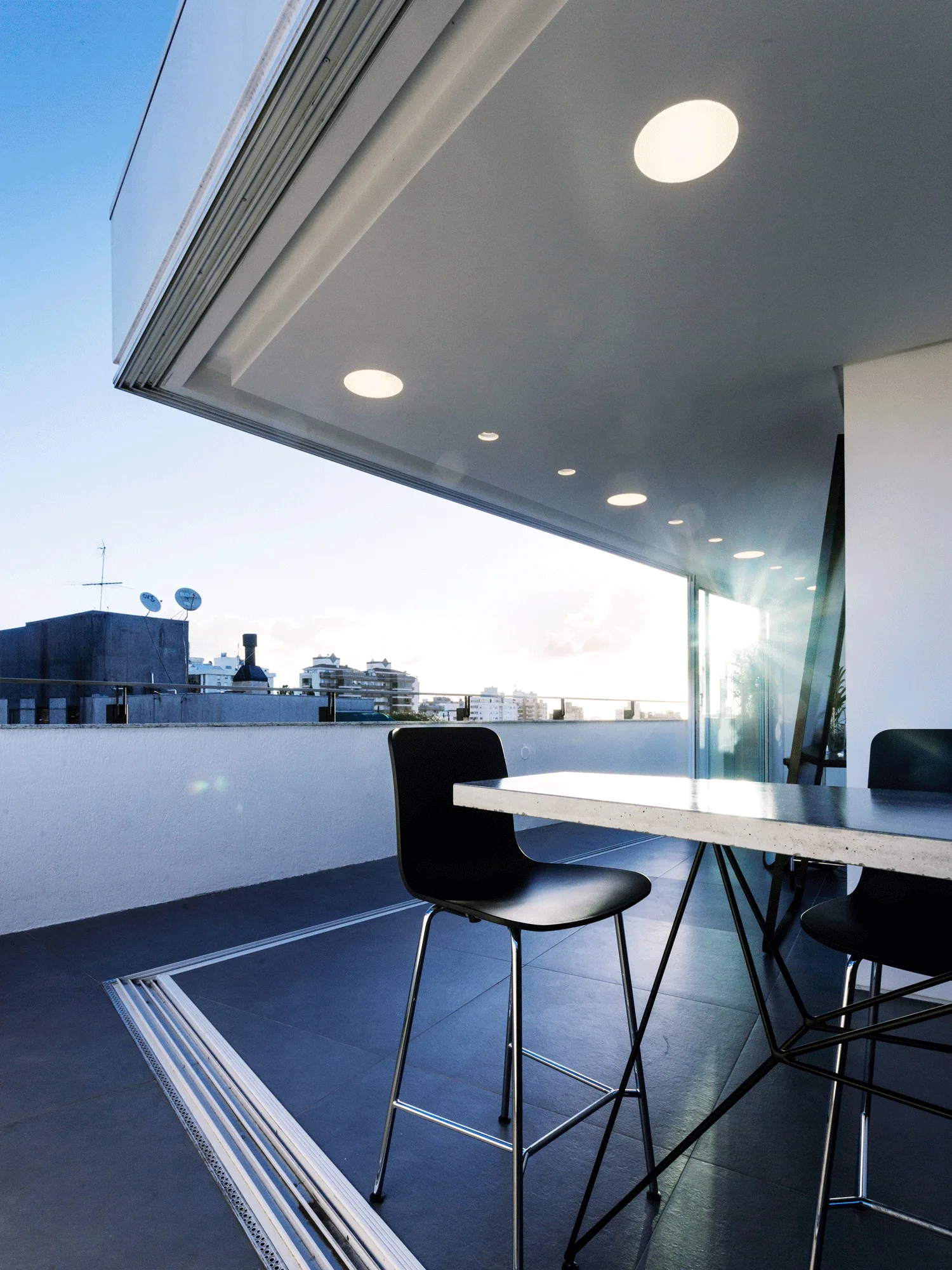 A modern high-rise balcony features a stone-topped bar table and black stools under a white ceiling with recessed lights looking out at a city skyline.