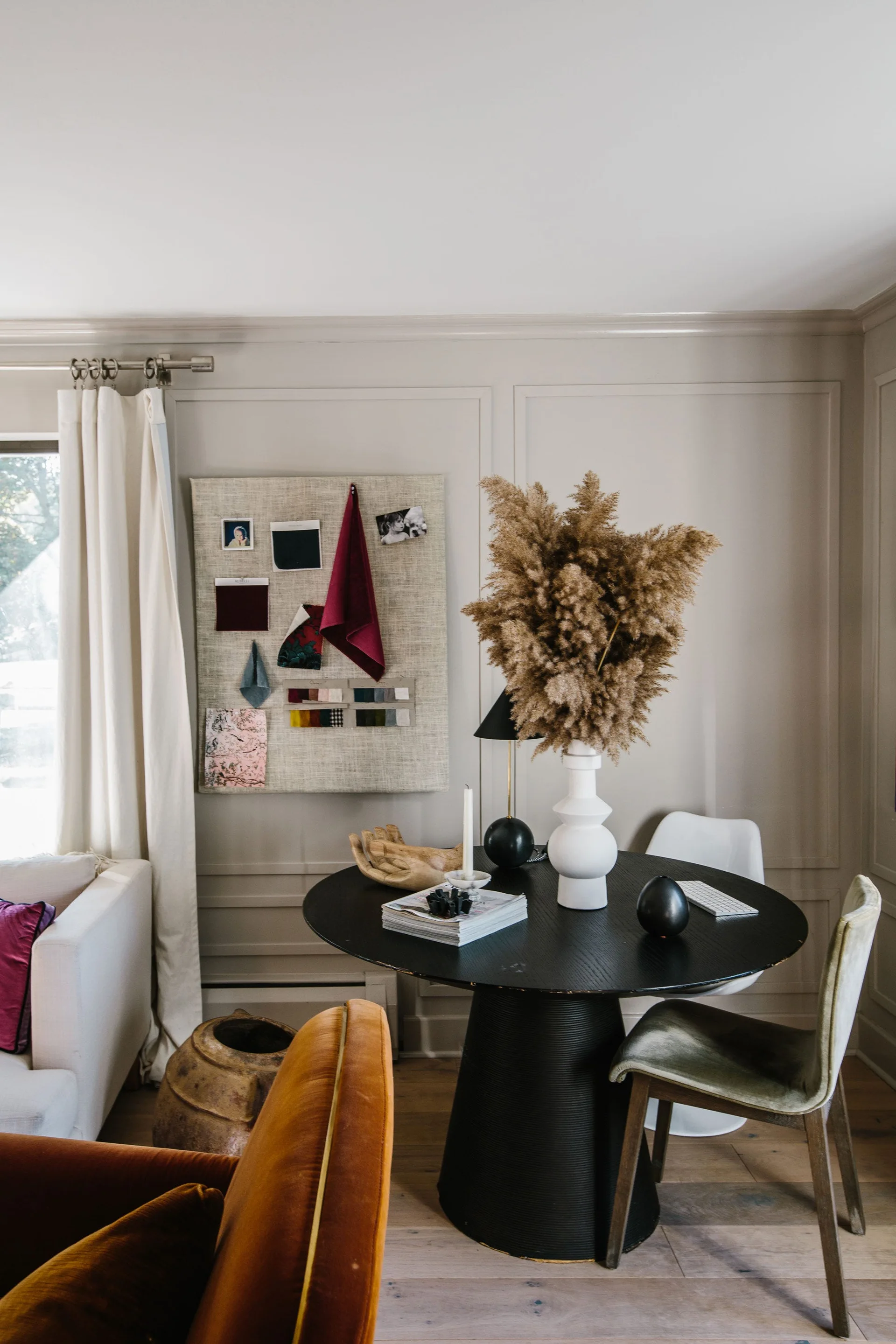 A round black dining table with a sculptural white vase and dried grass sits next to a beige paneled wall featuring a fabric mood board.