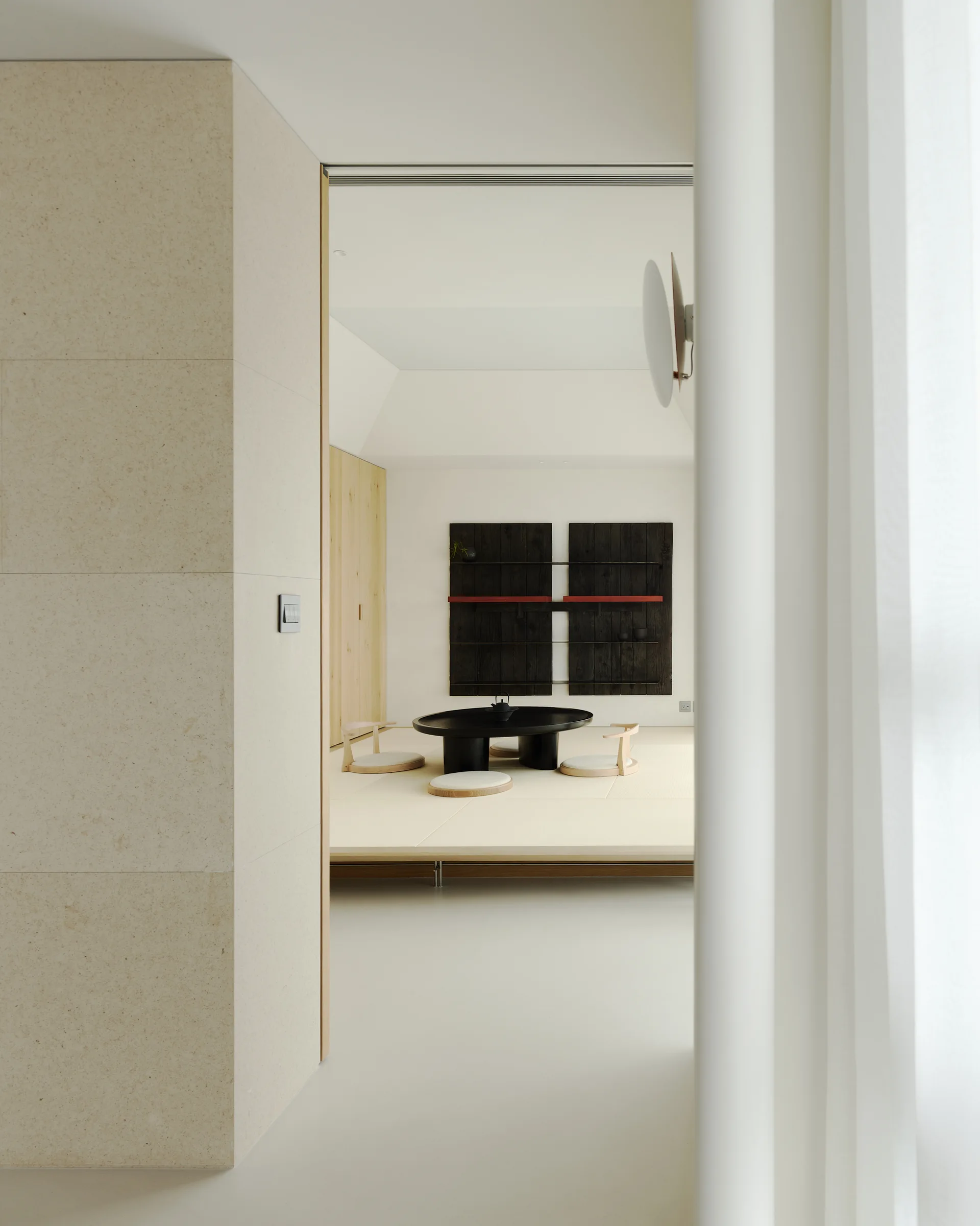A minimalist tatami room features a raised platform, a low black circular table with floor chairs, and a dark wall-mounted display shelf.