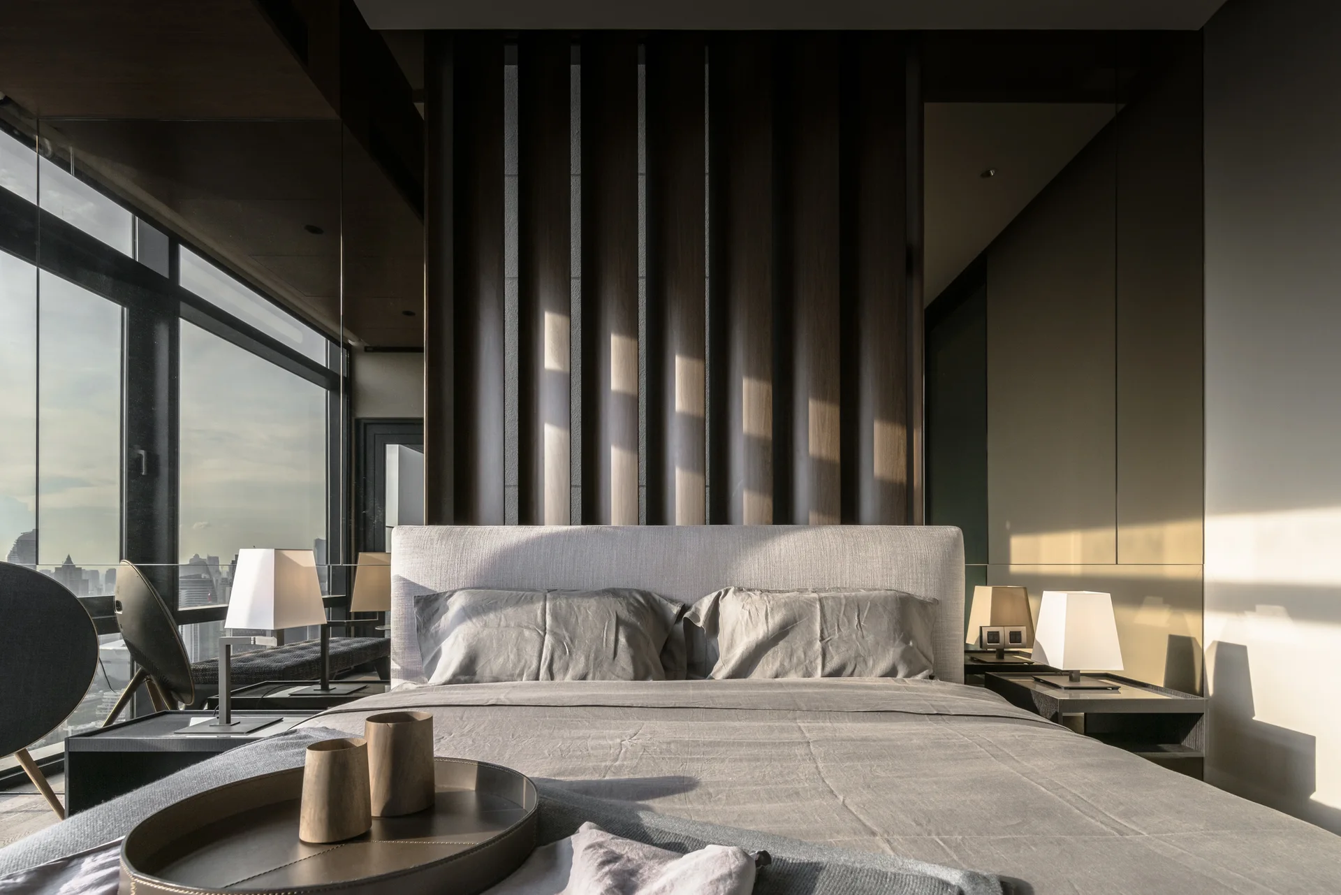 A contemporary bedroom features an upholstered grey bed against dark wood vertical wall panels next to a large window overlooking a city.