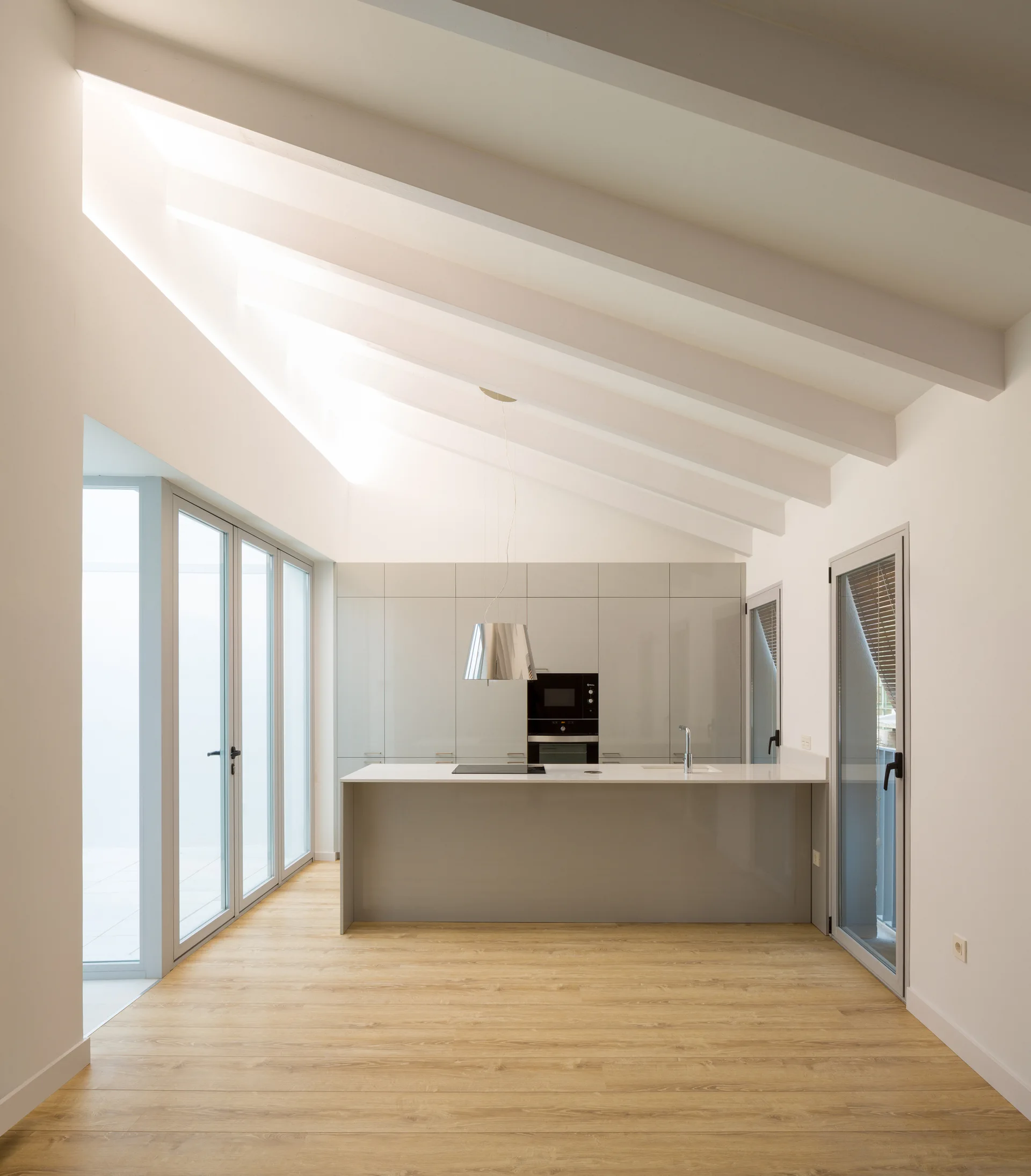 A minimalist kitchen features a grey central island and gloss cabinetry on light wood floors beneath a high vaulted ceiling with white beams.