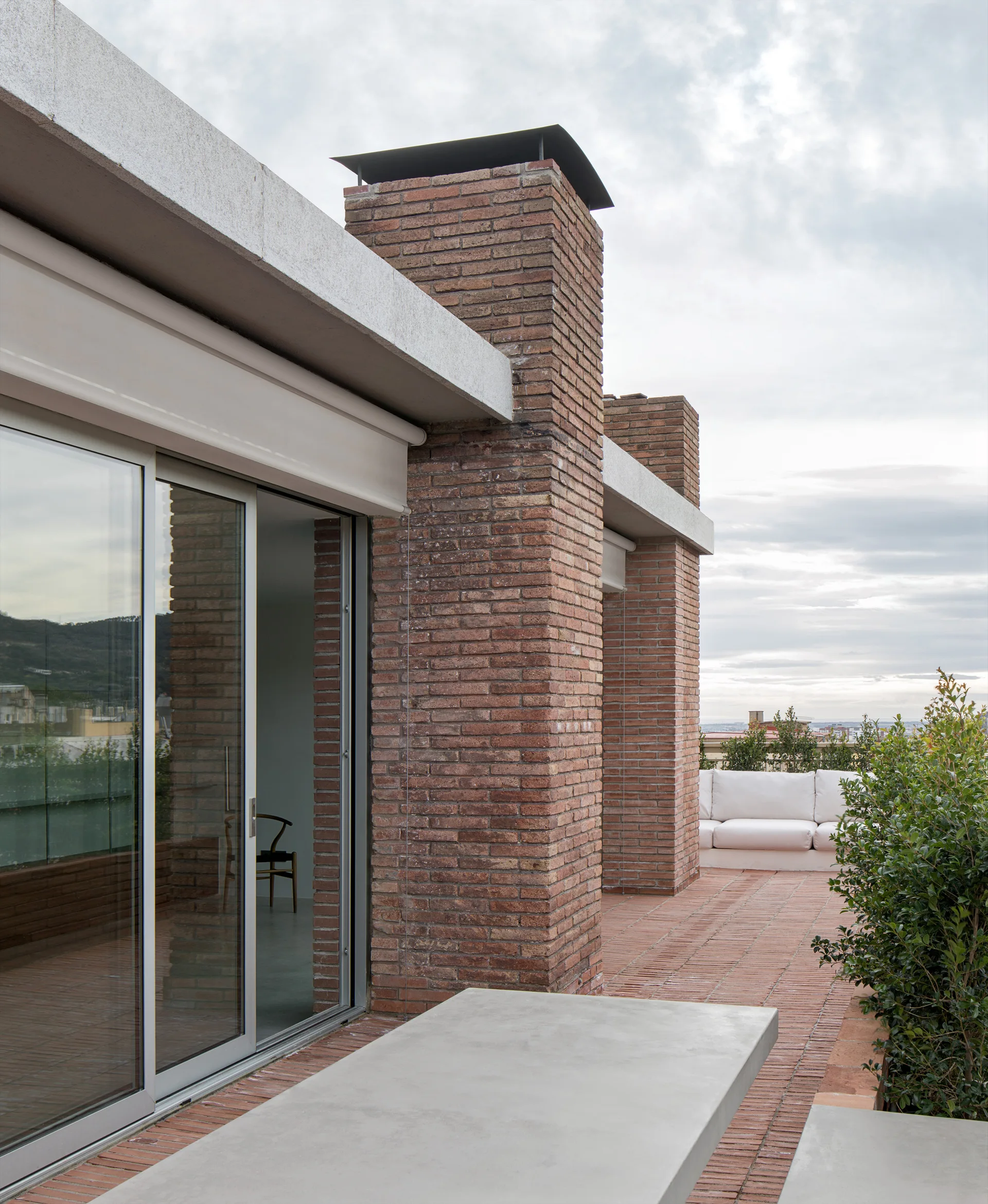 A modern rooftop terrace with red brick flooring, a large brick chimney, a sliding glass door, and a concrete dining table.