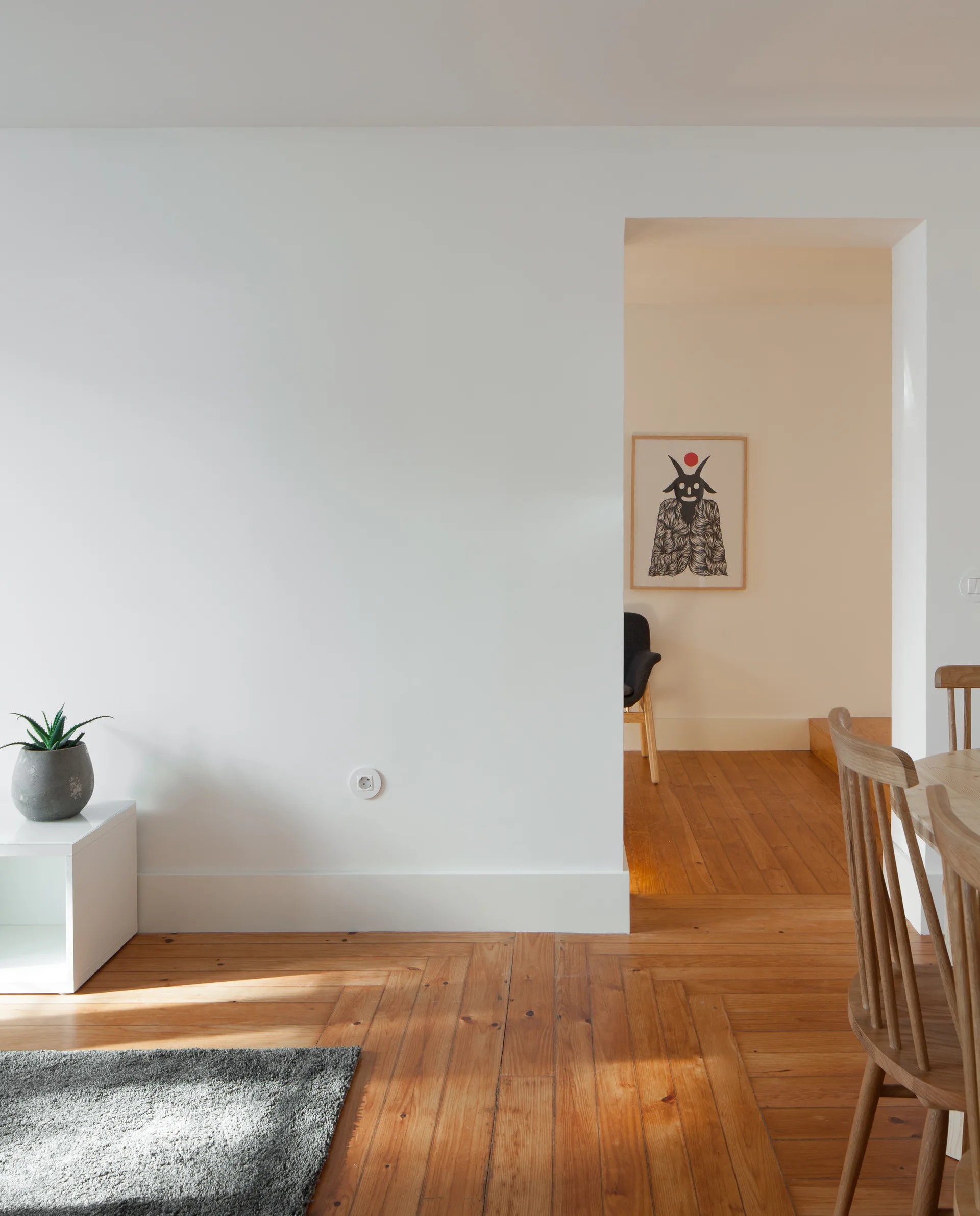 A minimalist dining room features a warm herringbone wood floor, a white wall with an open doorway, and wooden spindle dining chairs.