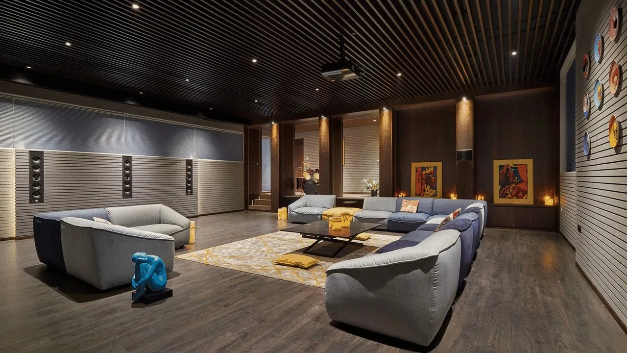 A large media room features modular grey sofas, a dark slatted acoustic ceiling, and wall-mounted speakers around a geometric pattern rug.