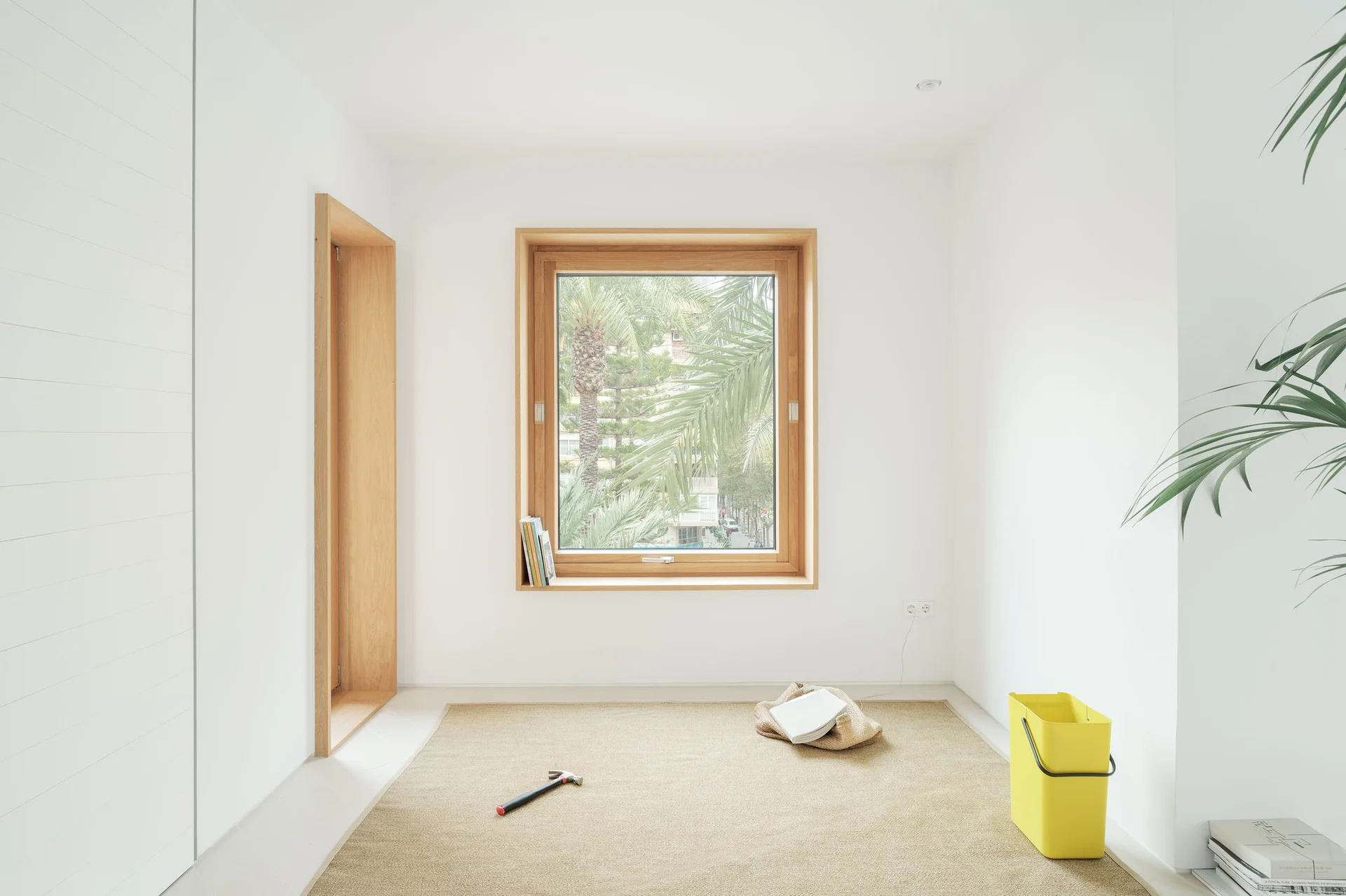 Bright minimalist interior with white walls, a deep wood-framed window, a matching wood doorway frame, and a beige woven rug.