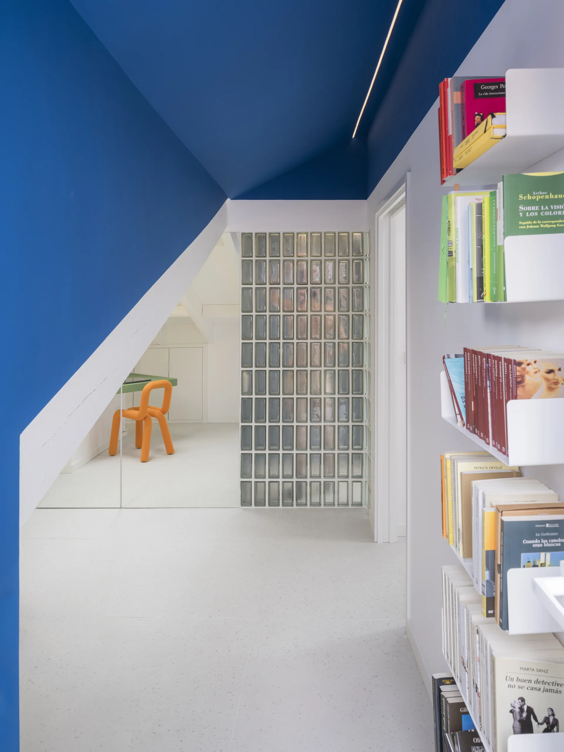 A modern hallway features a bold blue painted ceiling with linear lighting, white walls, built-in bookshelves, and a glass block divider.