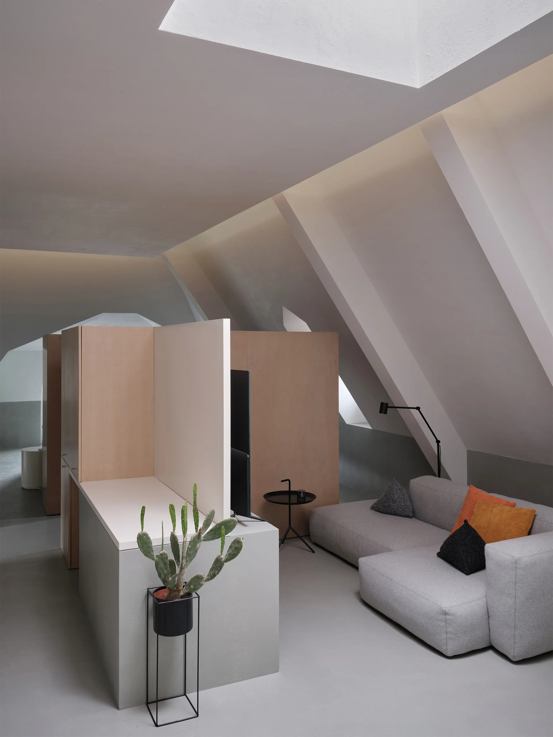 A modern attic living room featuring a gray modular sofa, a light wood partition, a slanted ceiling with a skylight, and a potted cactus.
