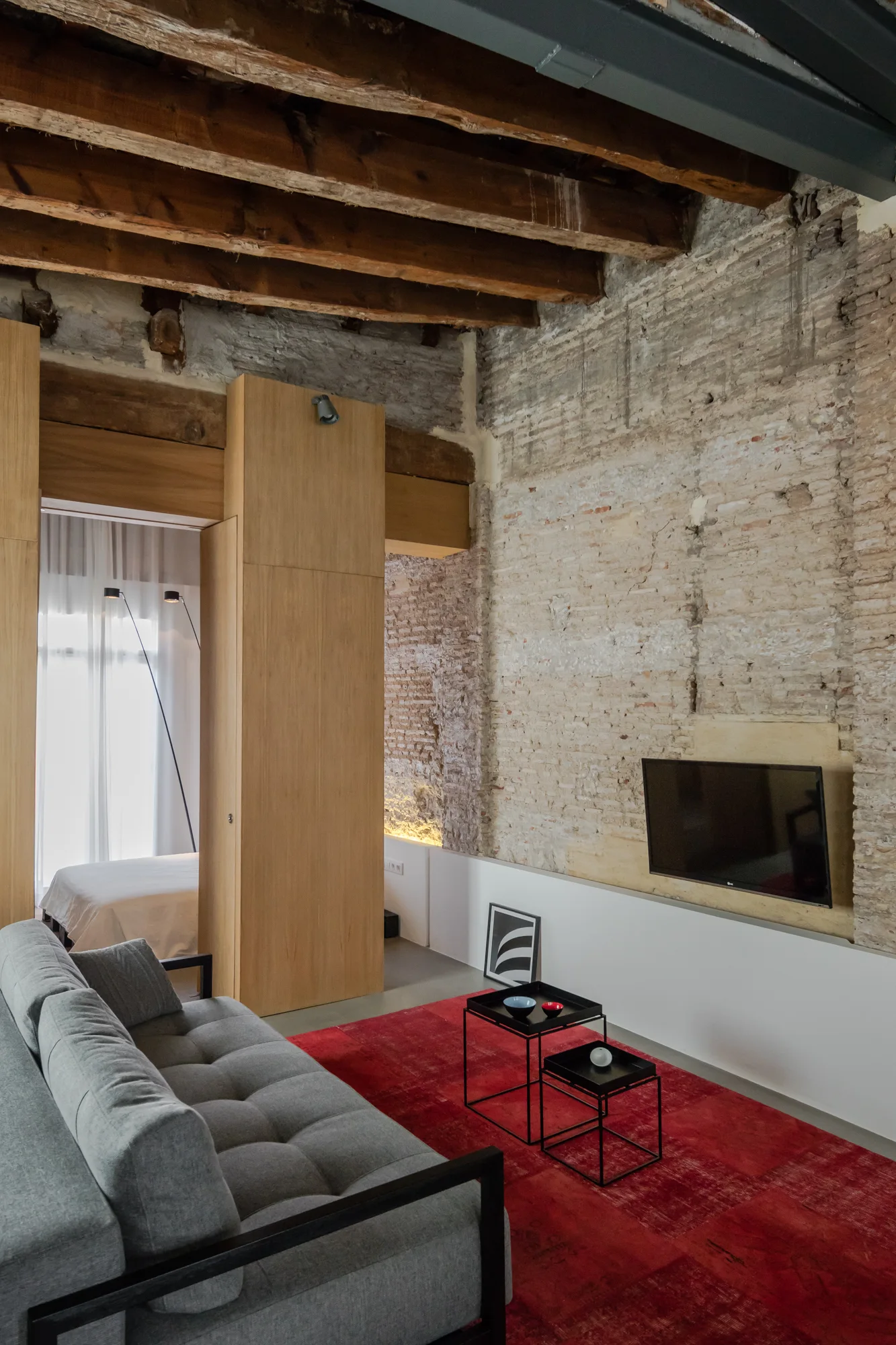 An industrial loft living room features a grey tufted sofa, a vibrant red rug, nesting coffee tables, and an exposed brick wall beneath wood ceiling beams.