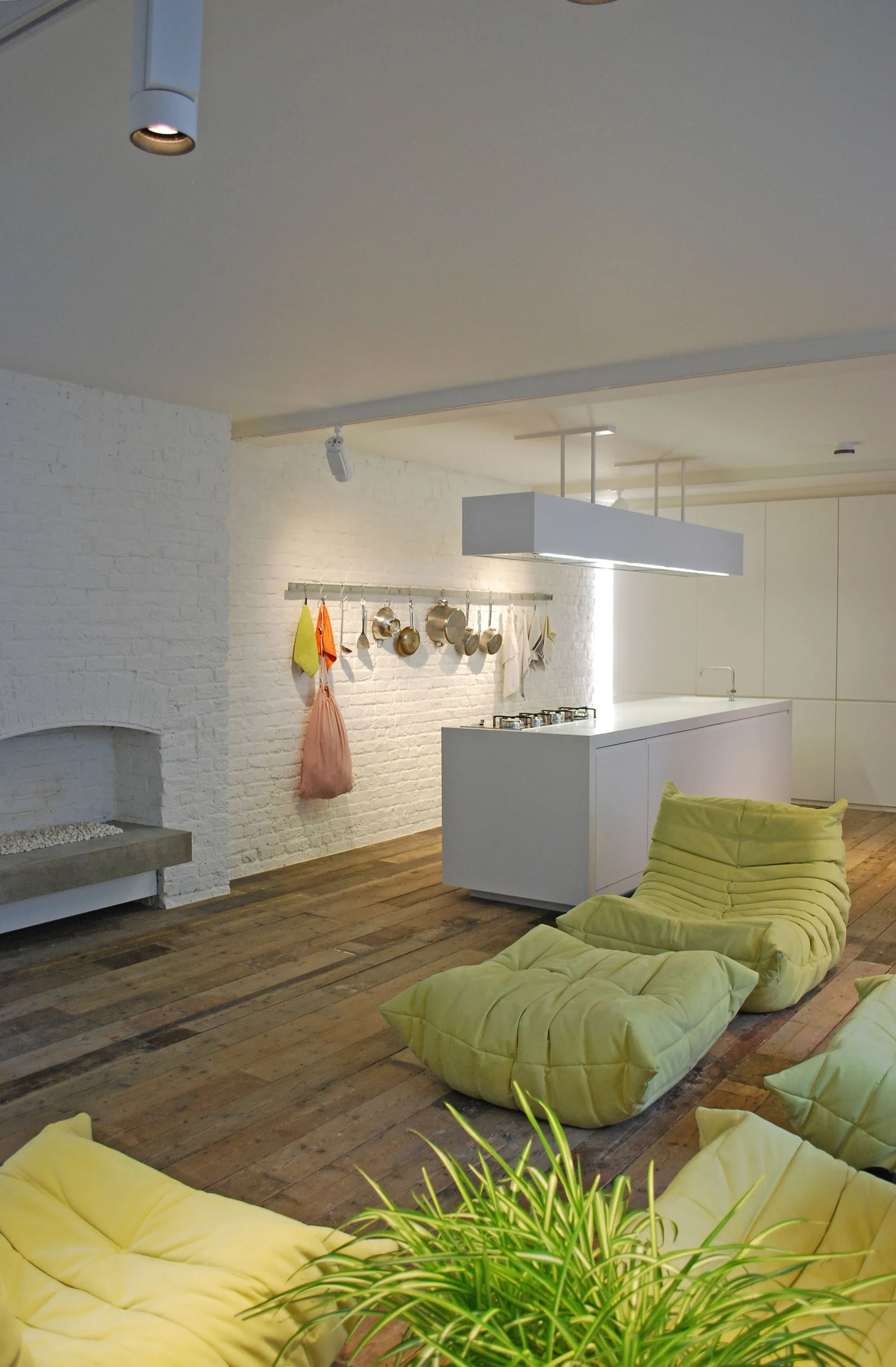 An open-concept kitchen features a white island, painted brick walls, reclaimed wood floors, and low-profile green modular chairs.