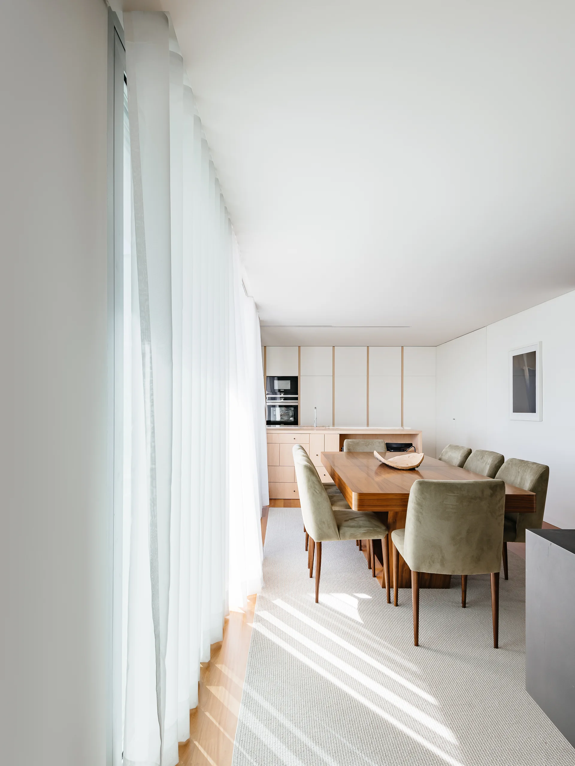 A modern dining room features a warm wood table, olive green upholstered chairs, a flat-woven rug, and floor-to-ceiling white sheer curtains.