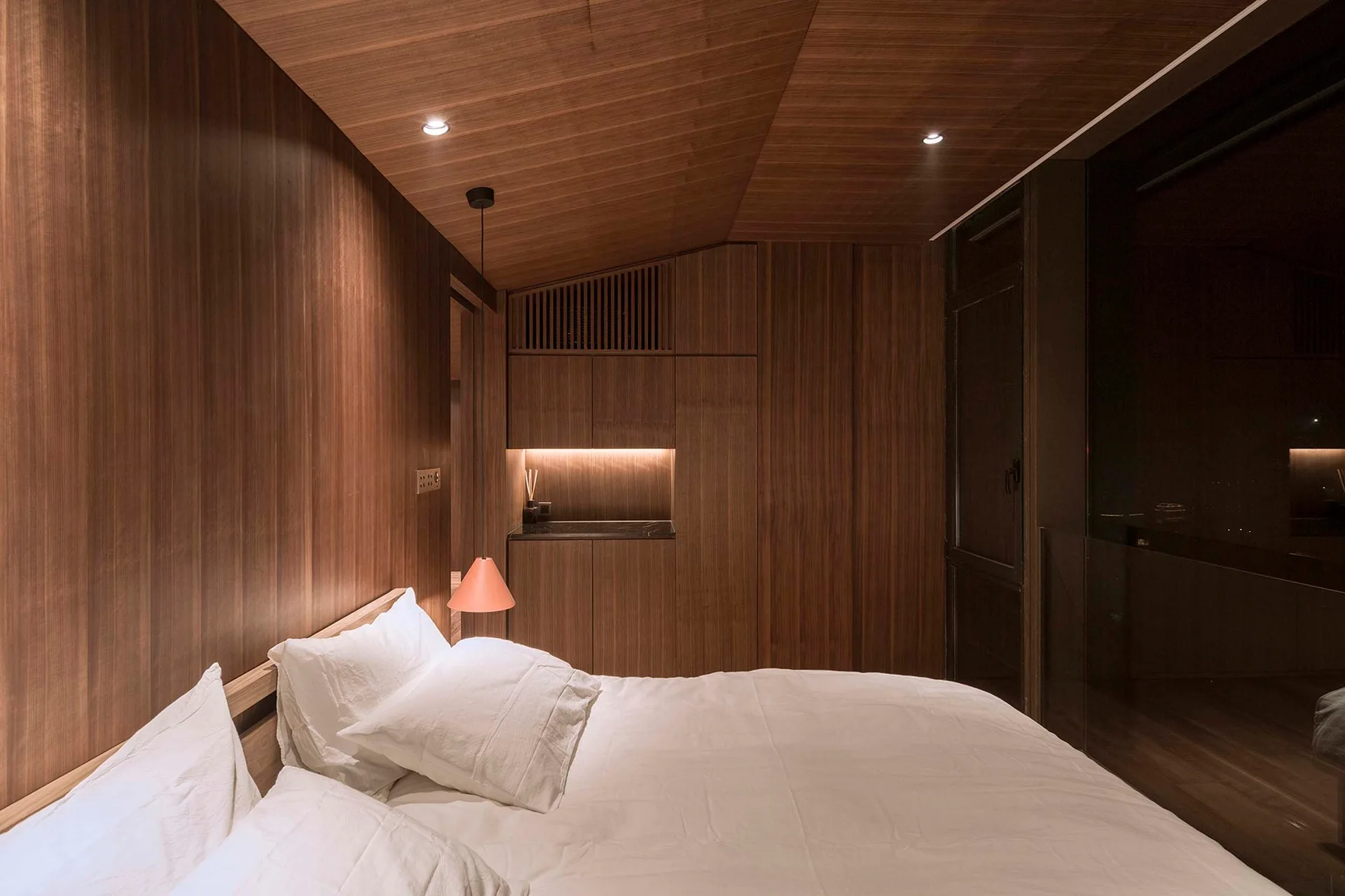 A bedroom features vertical wood paneling on the walls and ceiling, a bed with white sheets, and built-in matching cabinets with under-shelf lighting.