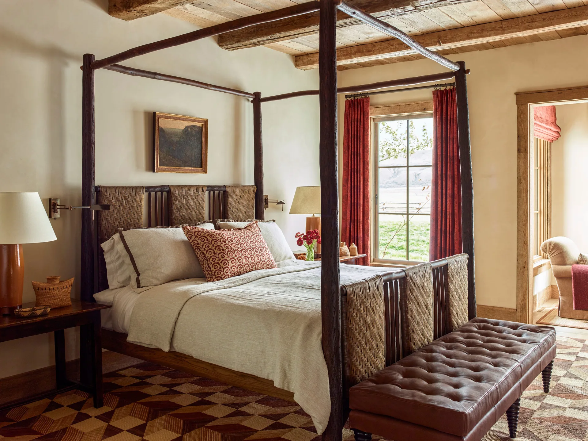 A rustic bedroom features a dark timber four-poster bed with woven headboards, a tufted leather bench, and a geometric patterned rug.