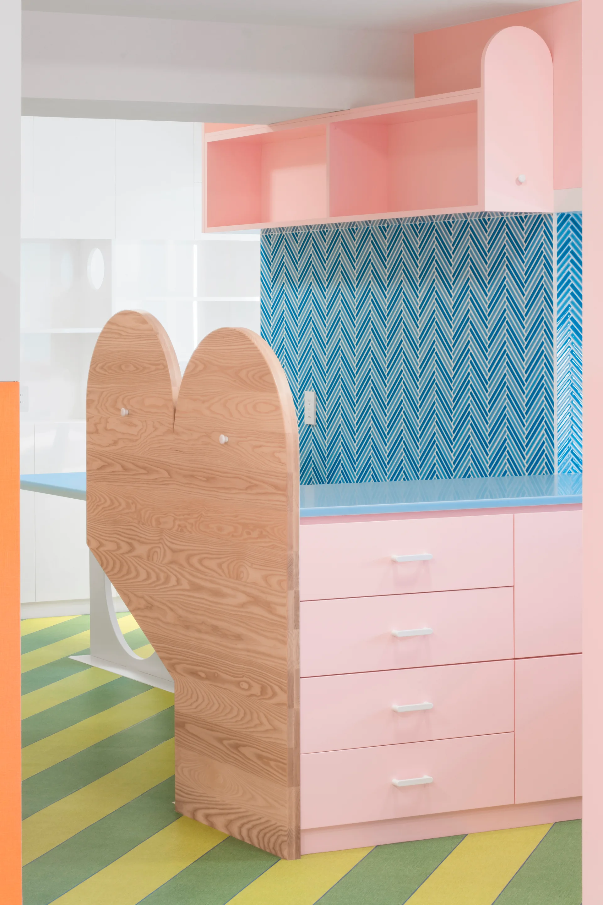 A pink playroom storage unit with a blue countertop, a heart-shaped wood side panel, and a blue herringbone tiled backsplash.