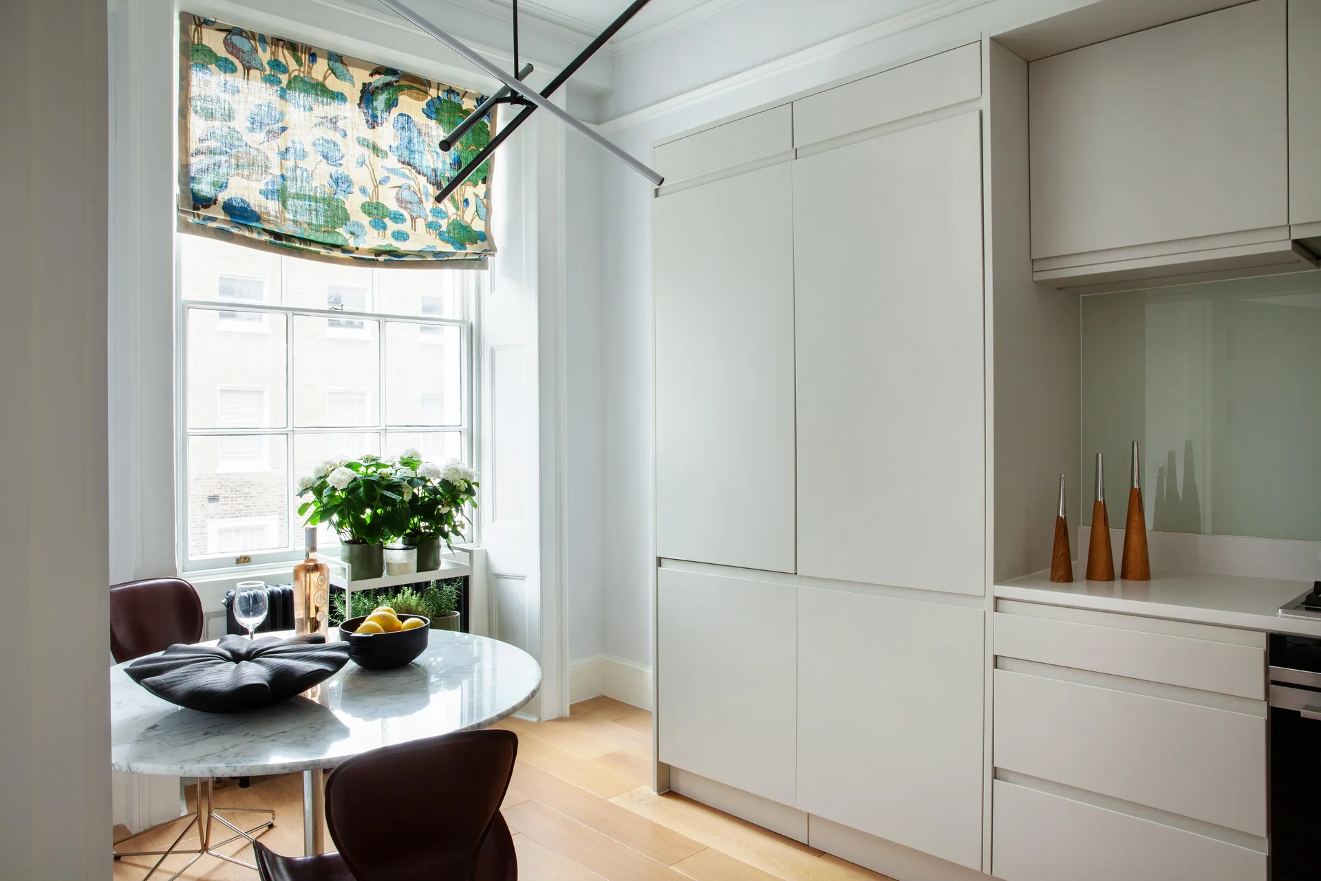 A light grey kitchen featuring tall matte cabinets, a round marble-top dining table with leather chairs, and a window with a patterned botanical shade.
