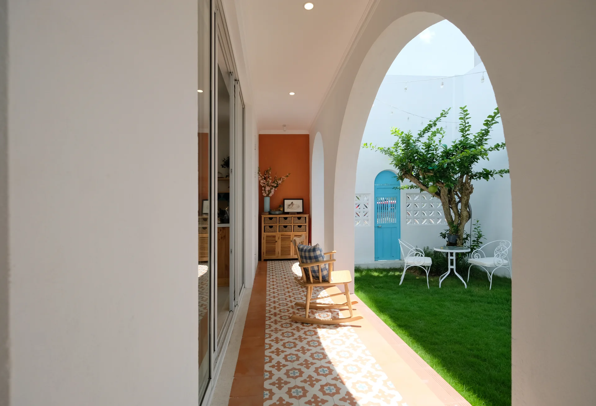 A covered white arched patio with patterned terracotta tiles and a wooden rocking chair looks out onto a green courtyard with a blue door.