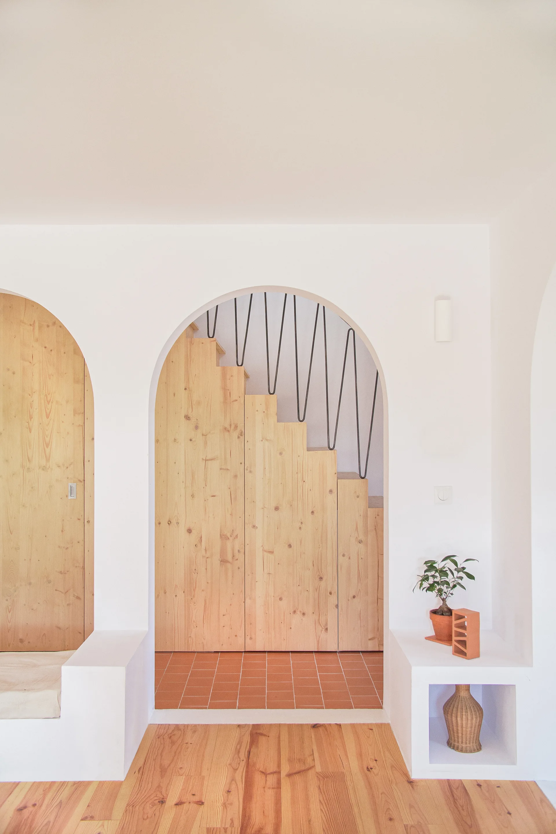 A white plastered wall with arched openings revealing a light wood staircase, terracotta floor tiles, and integrated display alcoves.