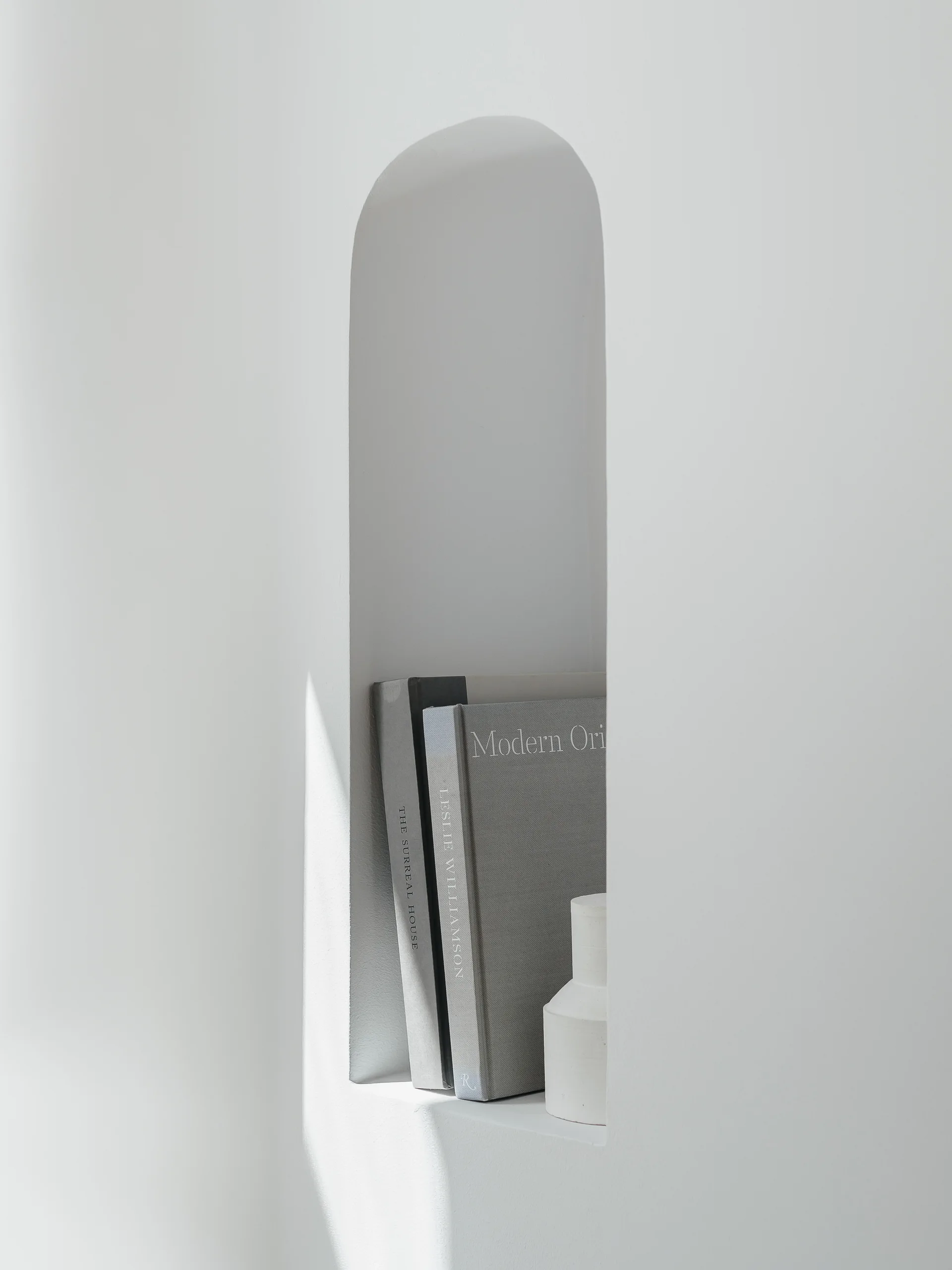A close-up of a minimalist recessed arched wall niche painted matte white, holding two gray books and a small white ceramic vase.