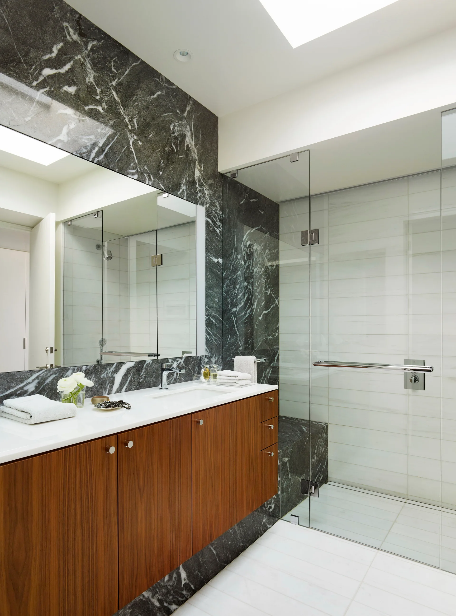 A modern bathroom features a floating wood vanity with a white countertop and a seamless glass shower against a dark veined marble wall.