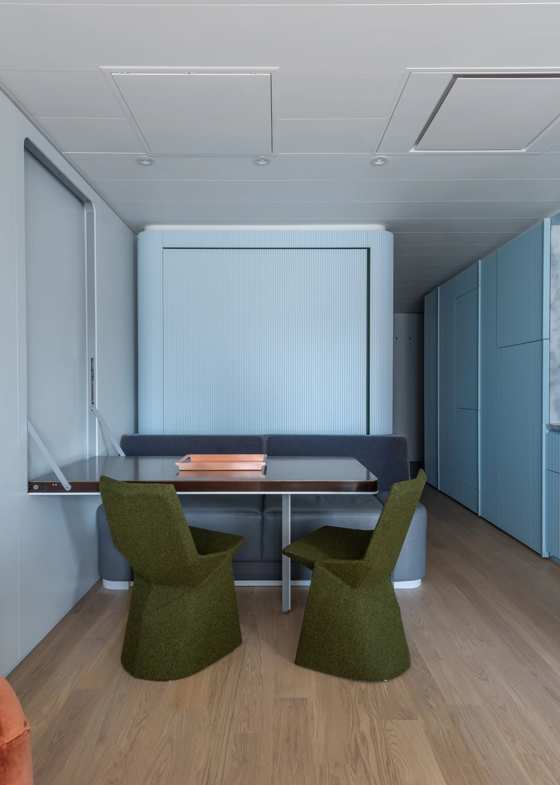 A modern dining nook features a rectangular table with two olive green chairs, a grey banquette, and light blue fluted wall paneling on wood flooring.