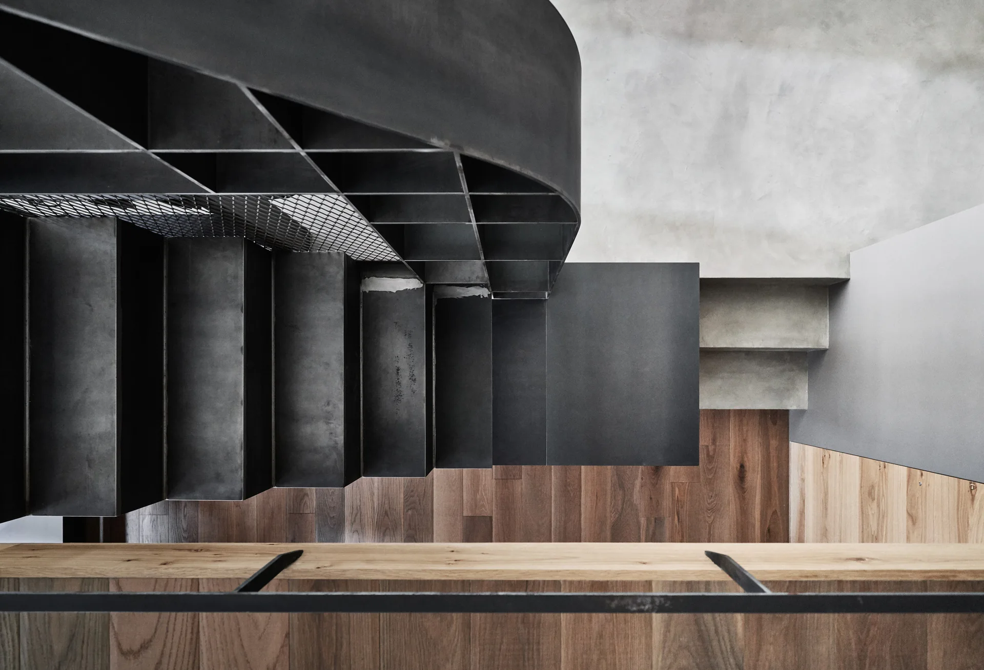 A top-down view of a dark metal staircase with a geometric grid railing next to a concrete landing and wood flooring below.
