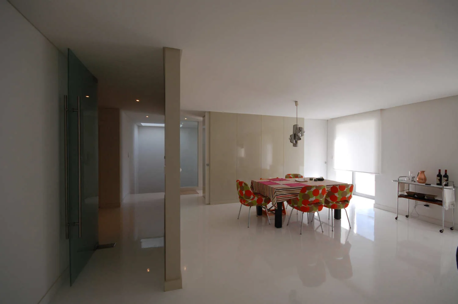 A minimalist dining room features a table with colorful patterned chairs on reflective glossy white floors next to a beige cabinet wall.