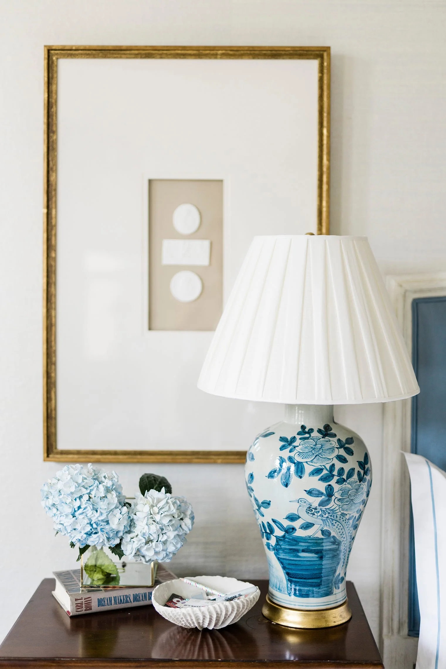 A dark wood bedside table with a blue and white porcelain lamp, fresh blue hydrangeas, and a gold-framed abstract artwork on the wall.