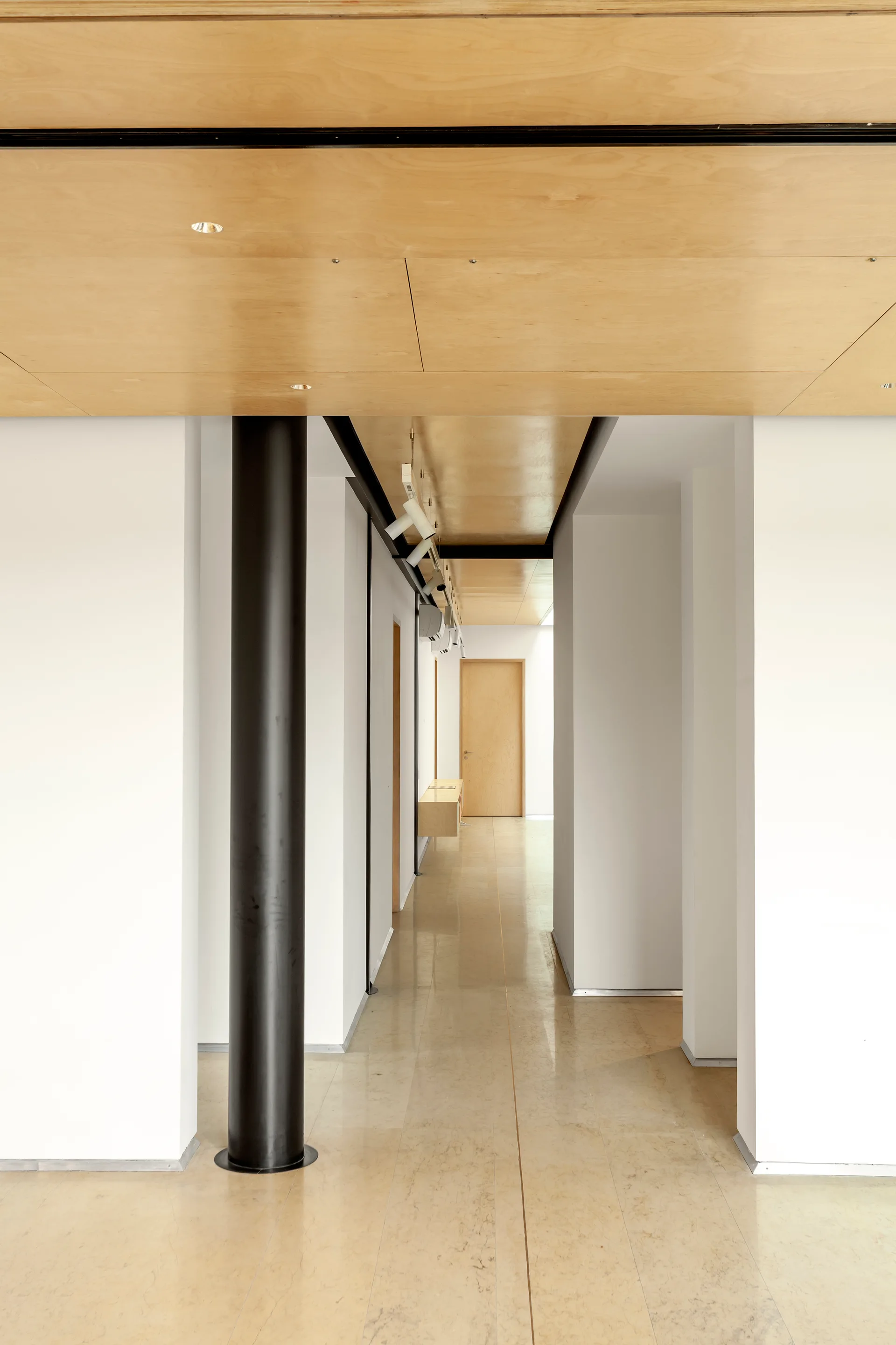 A minimalist hallway featuring white walls, light wood ceiling paneling, polished beige floors, and a prominent black structural column.