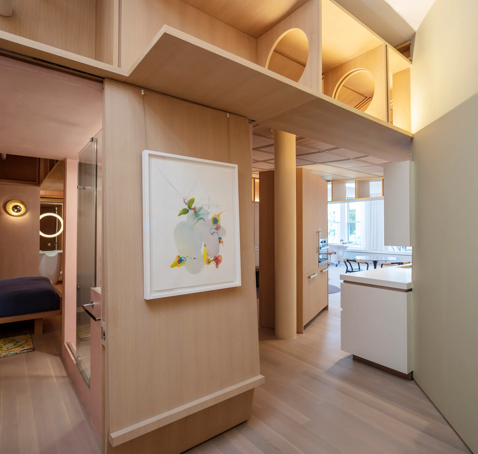 A modern residential interior features a light wood partition wall with framed art, overhead beams with circular cutouts, and a kitchen area.
