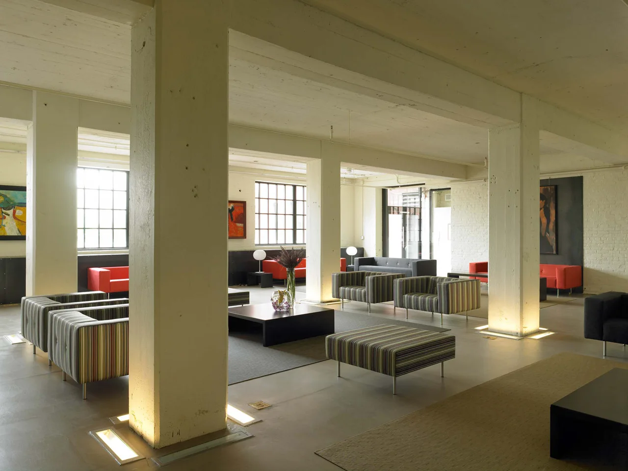 A spacious industrial loft lounge featuring large concrete structural columns with integrated floor uplighting, striped armchairs, and metal-framed windows.