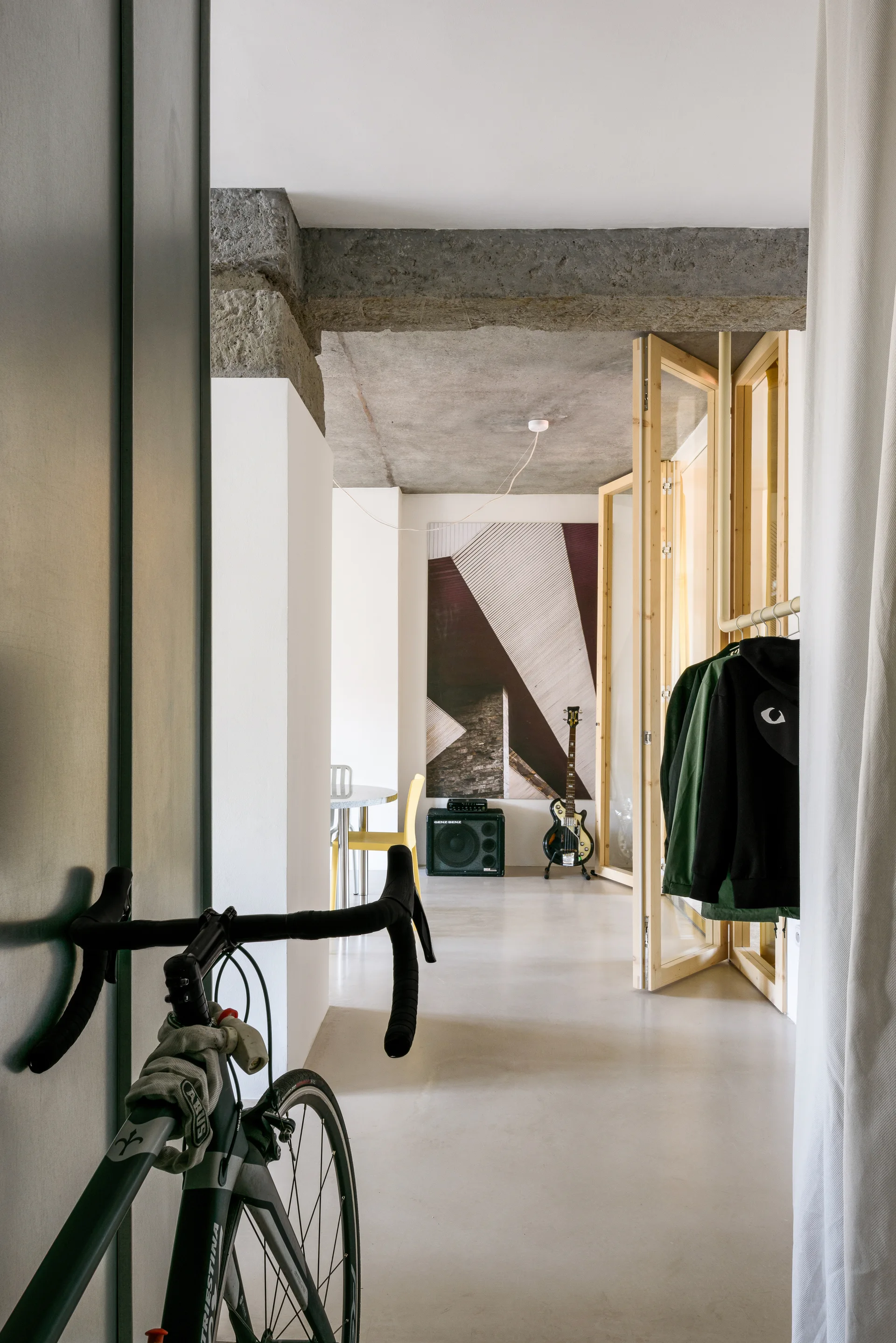 A loft entryway features a bicycle in the foreground, exposed concrete ceiling beams, a wooden folding door, and a hanging clothing rack.