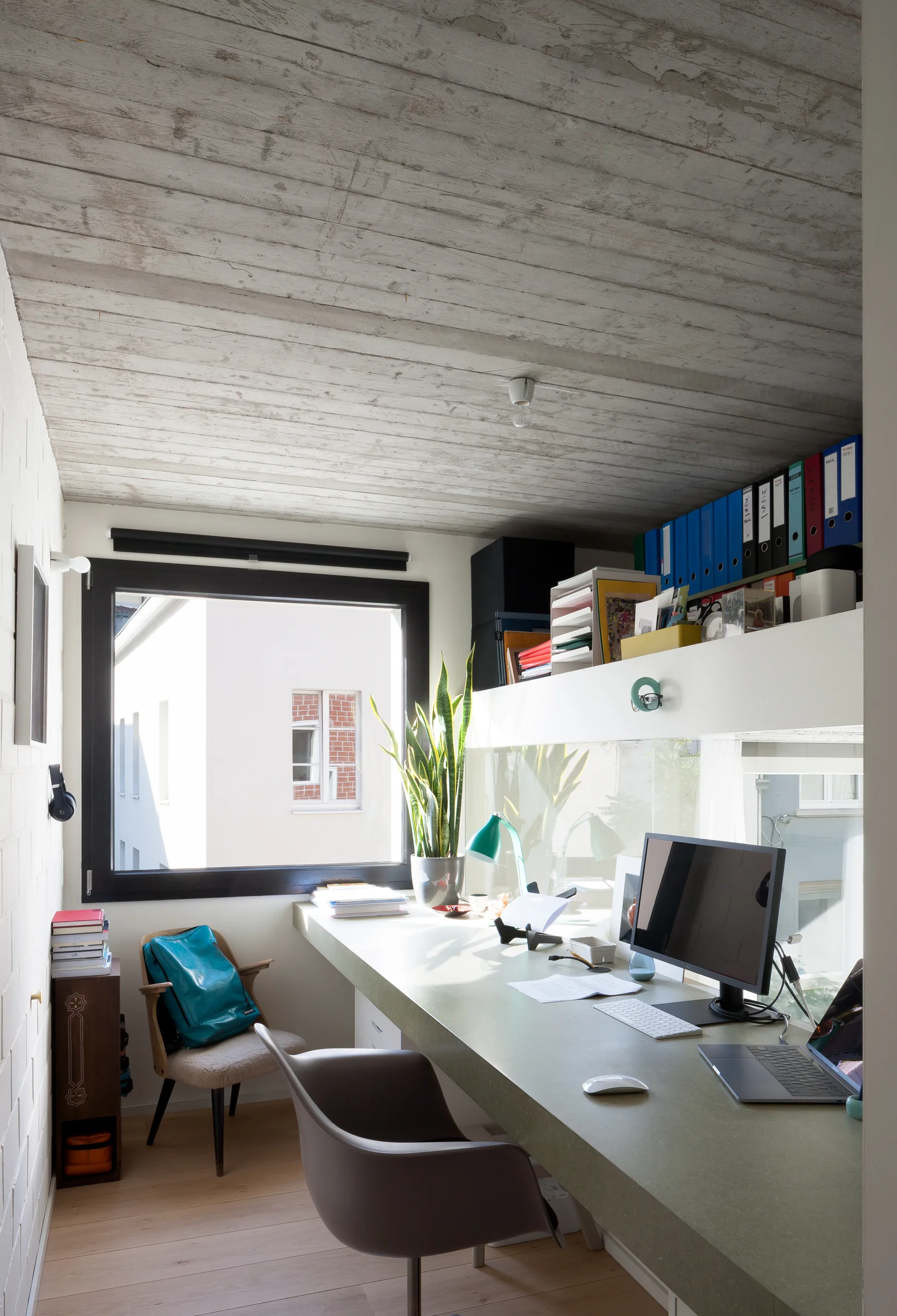 A compact home office features a long green built-in desk with a computer, a modern gray chair, and a high storage shelf under a textured wood ceiling.
