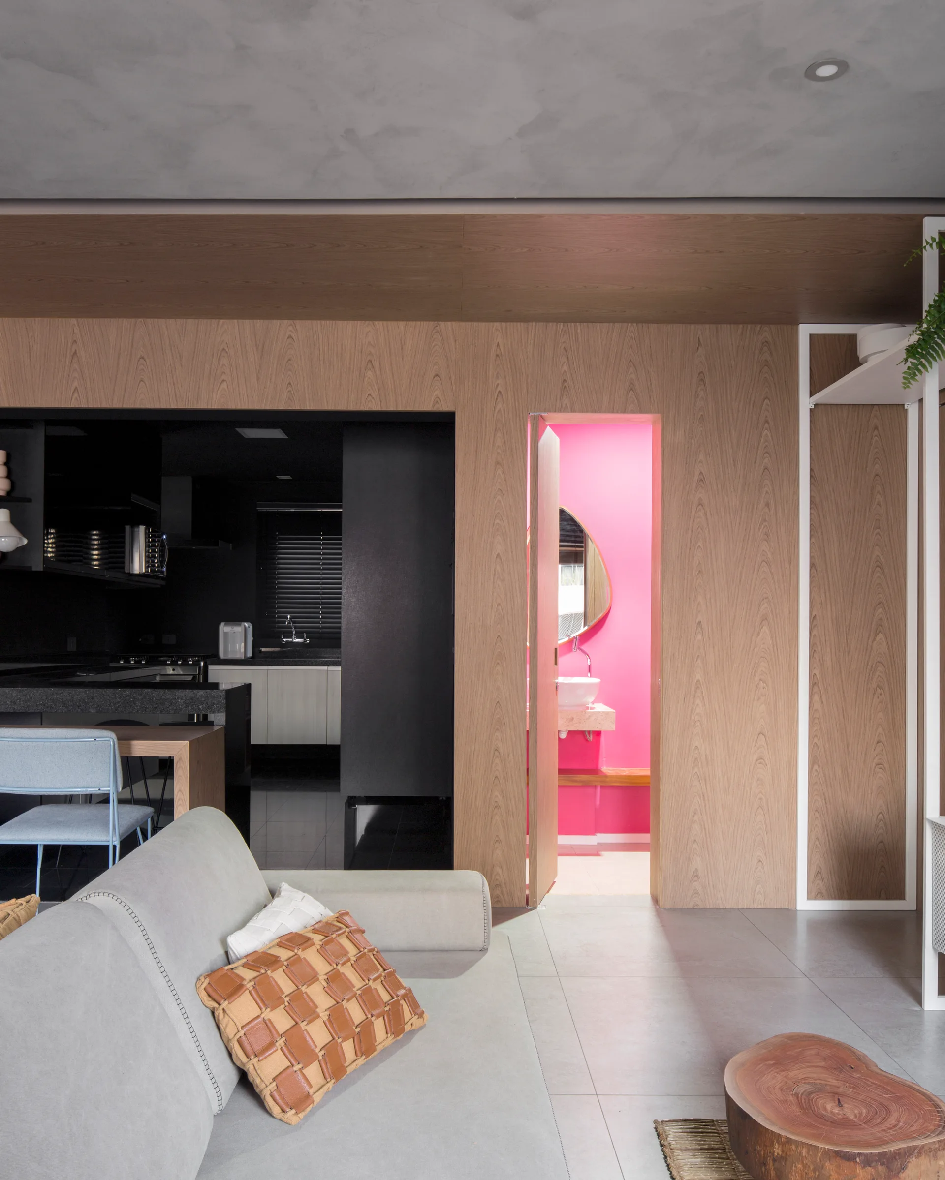 A grey sofa sits in a living room with wood-paneled walls, showing a doorway into a bright pink bathroom and a glimpse of a dark kitchen.