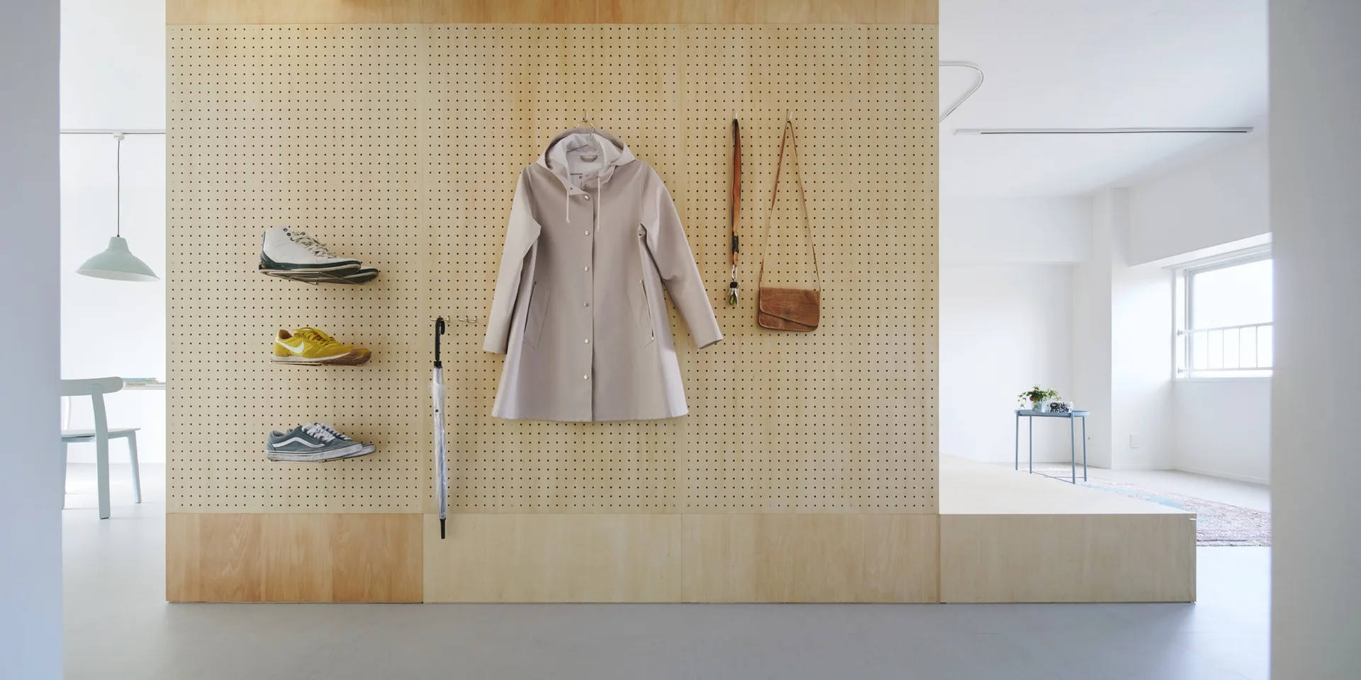 A wood pegboard wall organizes a hanging coat and shoes on shelves next to an elevated platform floor in a bright entryway.