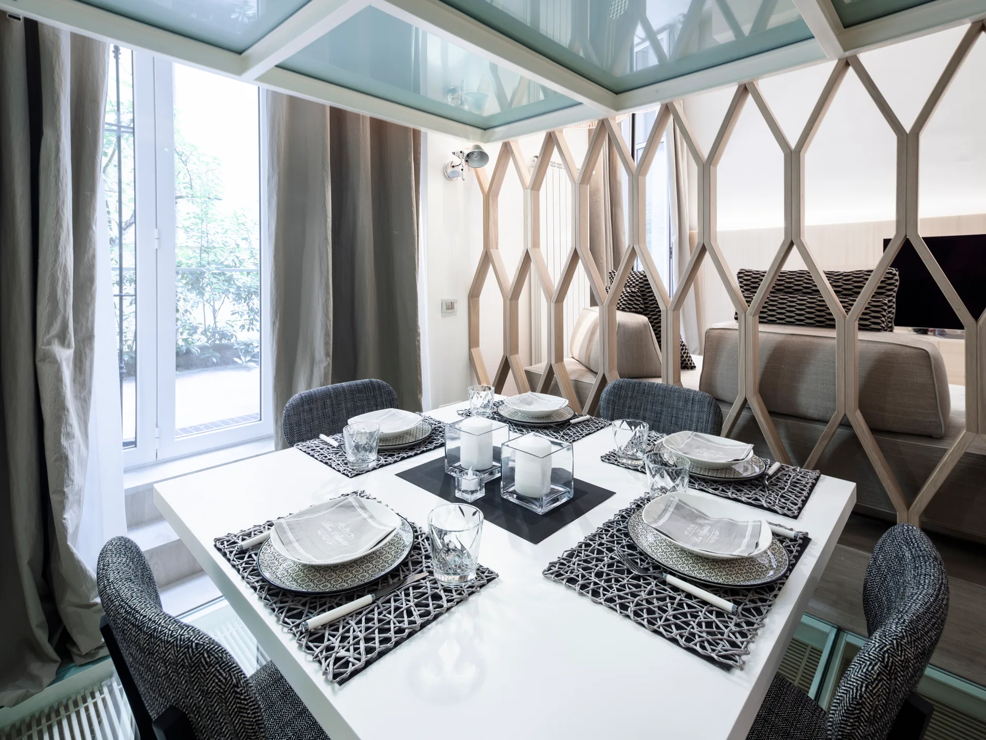 A modern white dining table set for four is positioned next to a geometric light wood screen partition in a bright room.