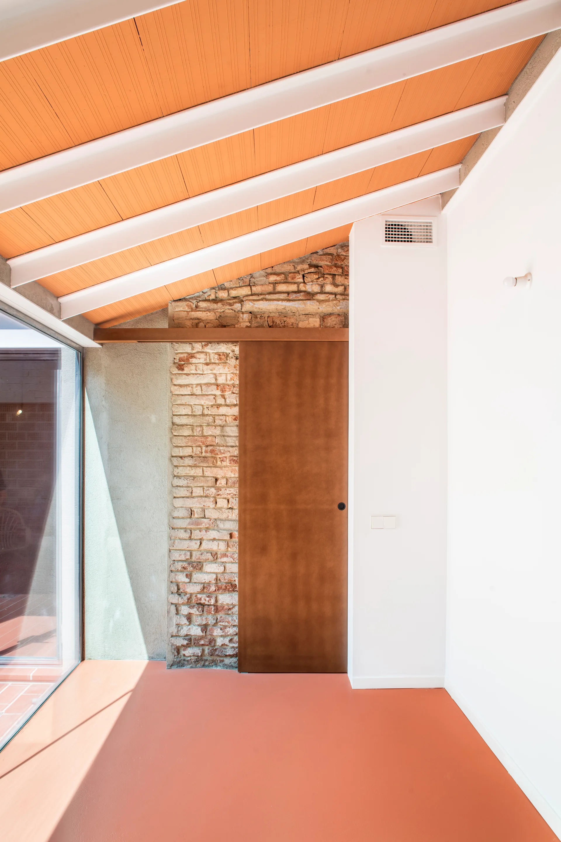 A modern room corner with a slanted terracotta ceiling, white support beams, an exposed brick wall, and a large brown sliding door.
