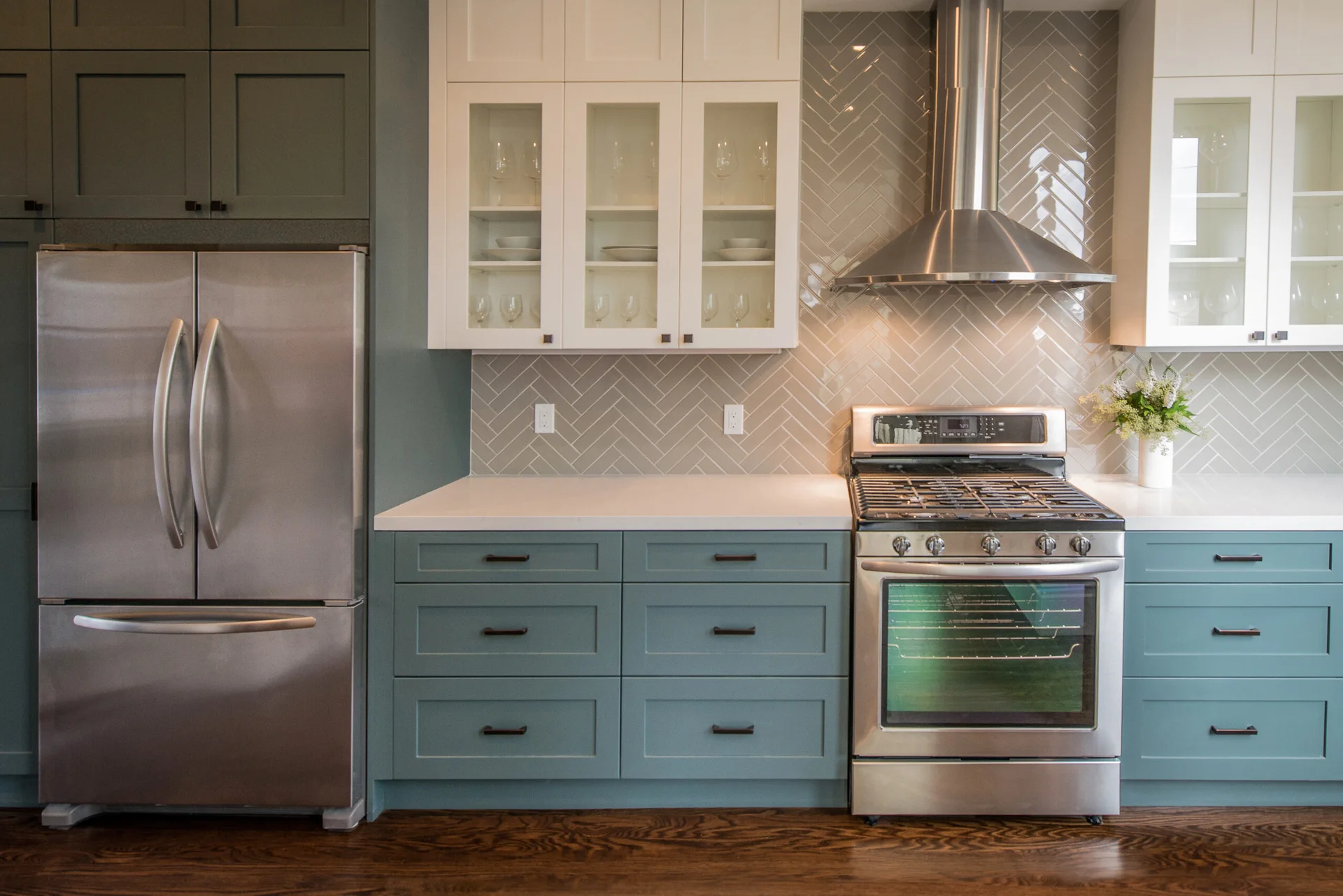 A kitchen features matte teal lower cabinets, white upper cabinets with glass panels, a glossy grey herringbone tile backsplash, and stainless steel appliances.