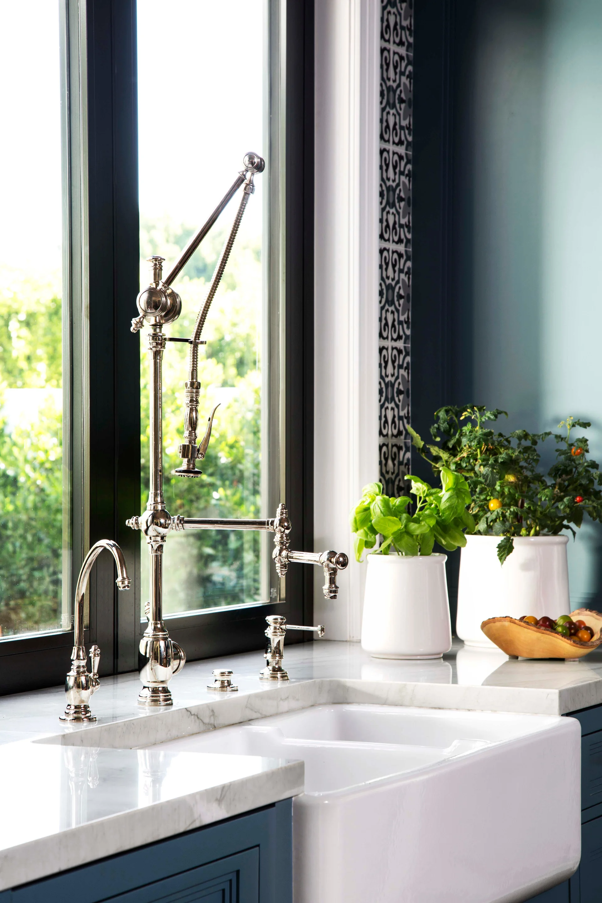 A polished nickel faucet sits on a marble countertop above a white farmhouse sink next to a black-framed window.