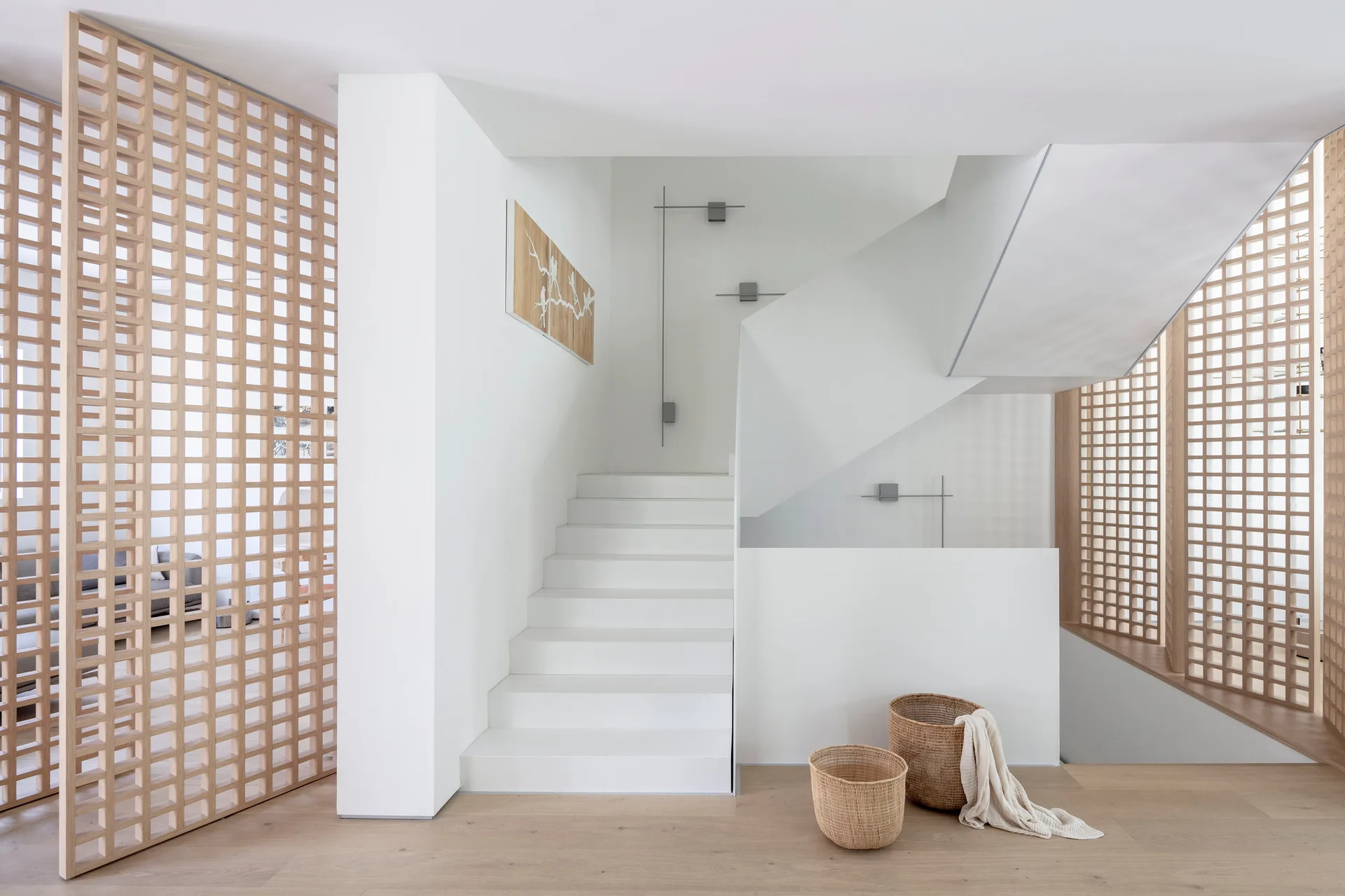 A minimalist white staircase is flanked by light wood grid room dividers and two woven baskets on a light wood floor.