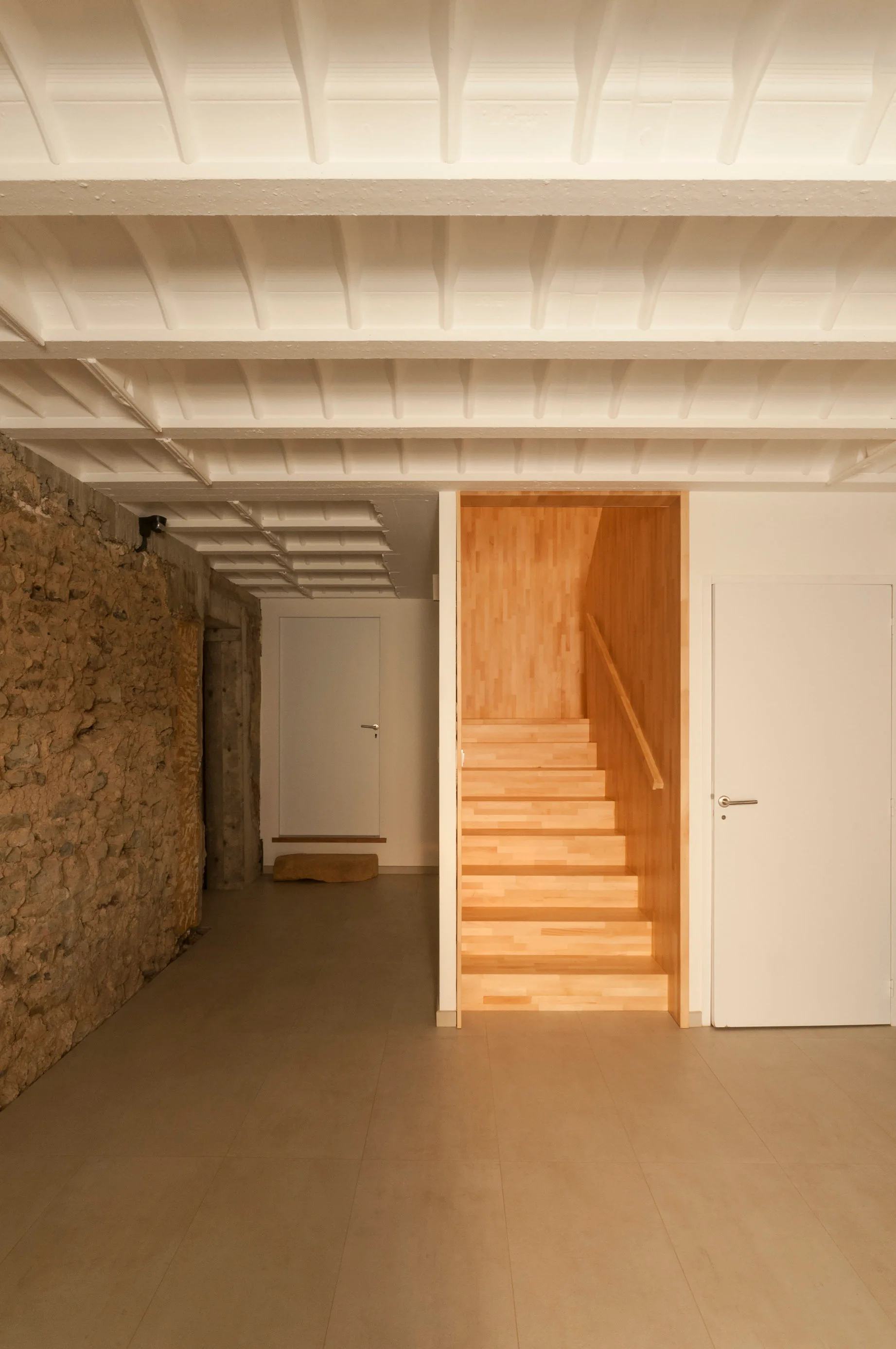A minimalist hallway features an enclosed light wood staircase, an exposed stone wall, a white coffered ceiling, and beige tile flooring.