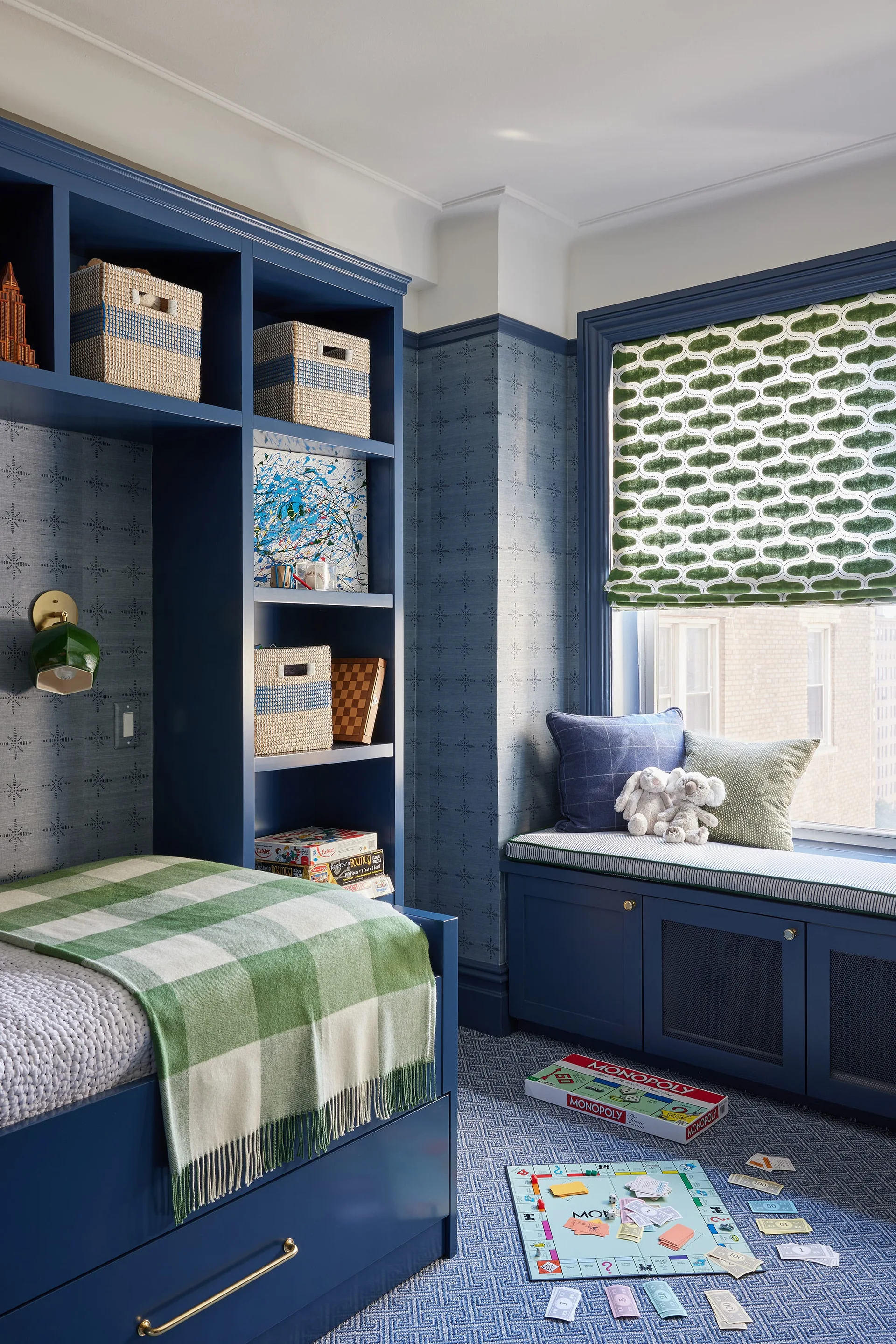 A children's bedroom features custom blue built-in shelving, a matching window seat, patterned wallpaper, and a green checked blanket.